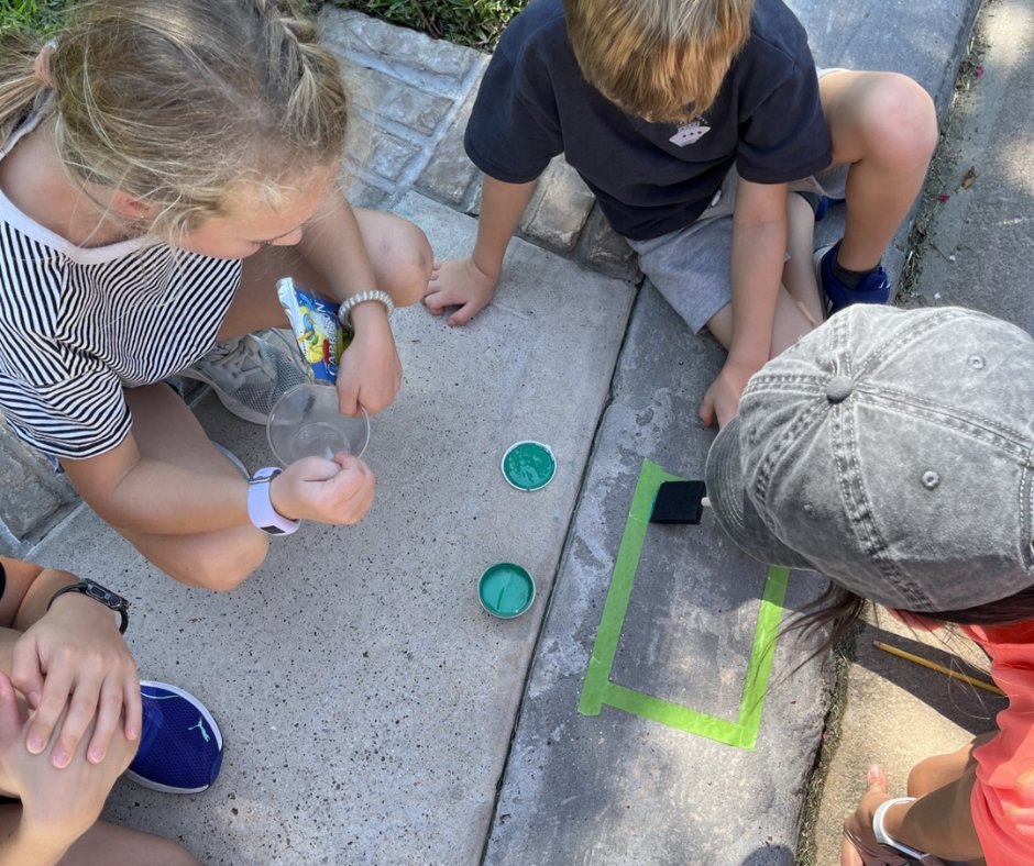 Children and an adult paint a stenciled address number on a concrete curb outdoors.