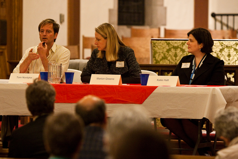 Three candidates seated at a table with name placards at an indoor candidate forum.