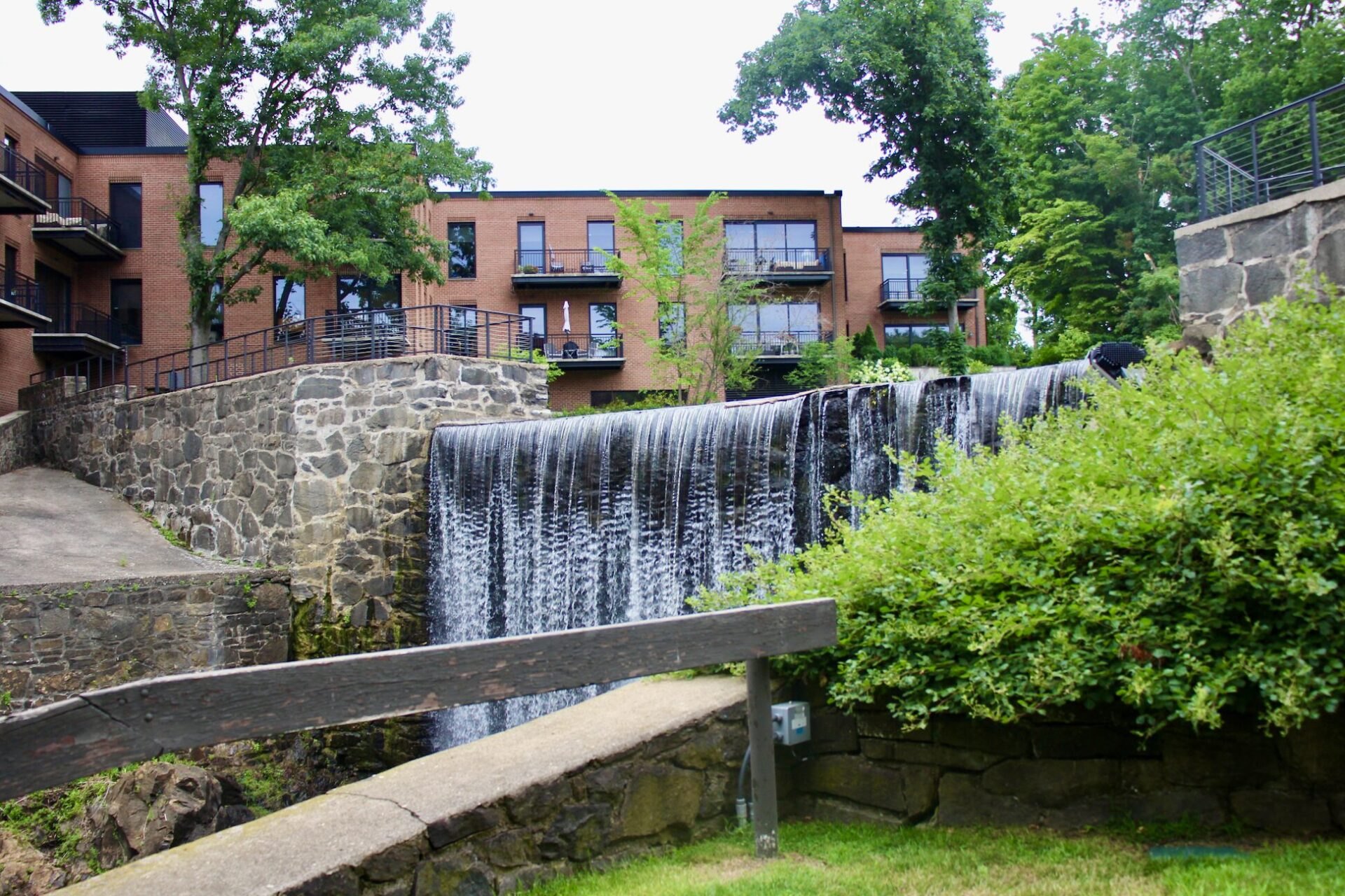 Waterfall cascading over a stone dam next to a multi-story brick building complex with balconies.