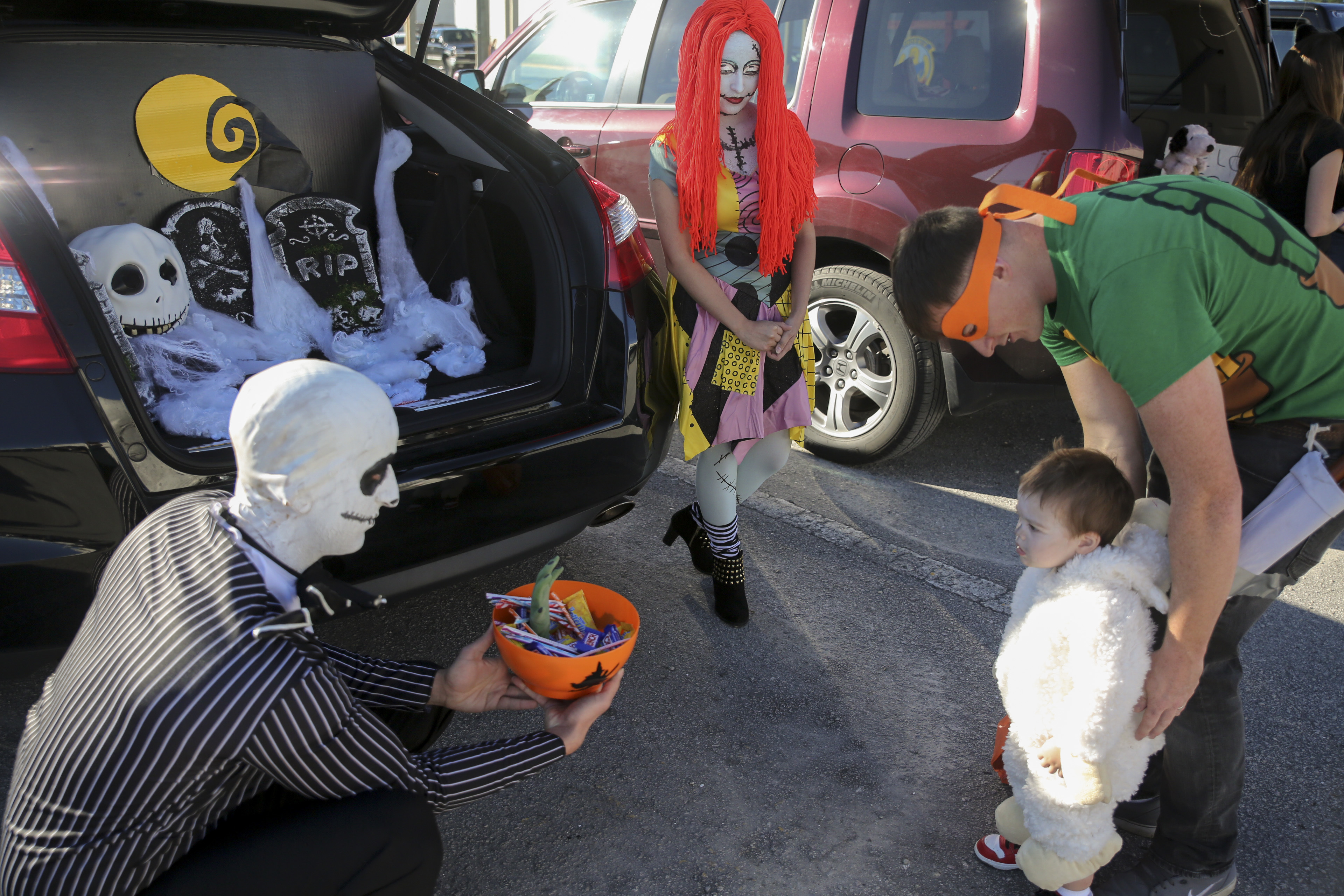 Costumed adults handing candy from a decorated car trunk to a small child at a Halloween event.