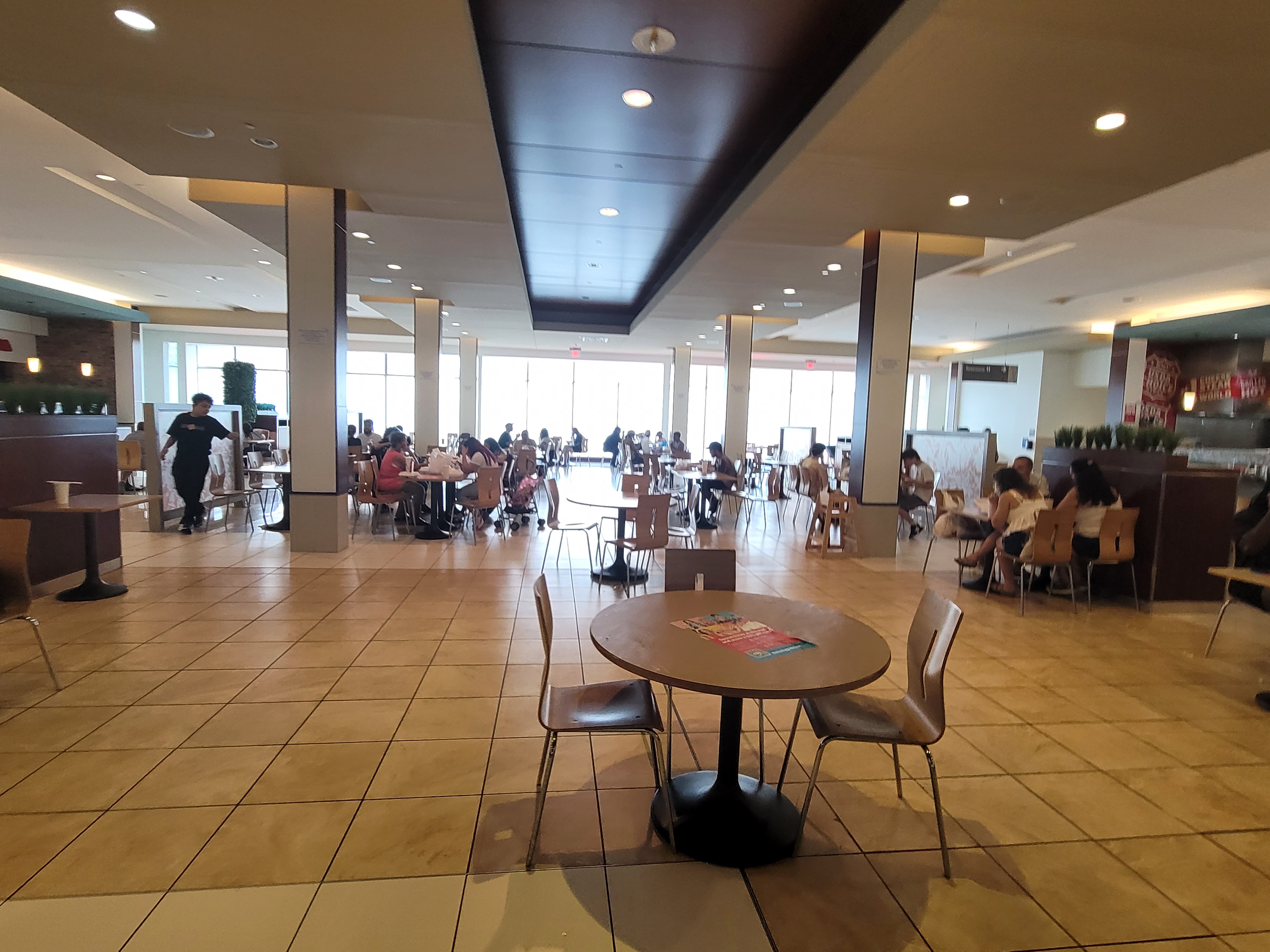 Spacious mall food court with tables, chairs, and diners under recessed lighting.