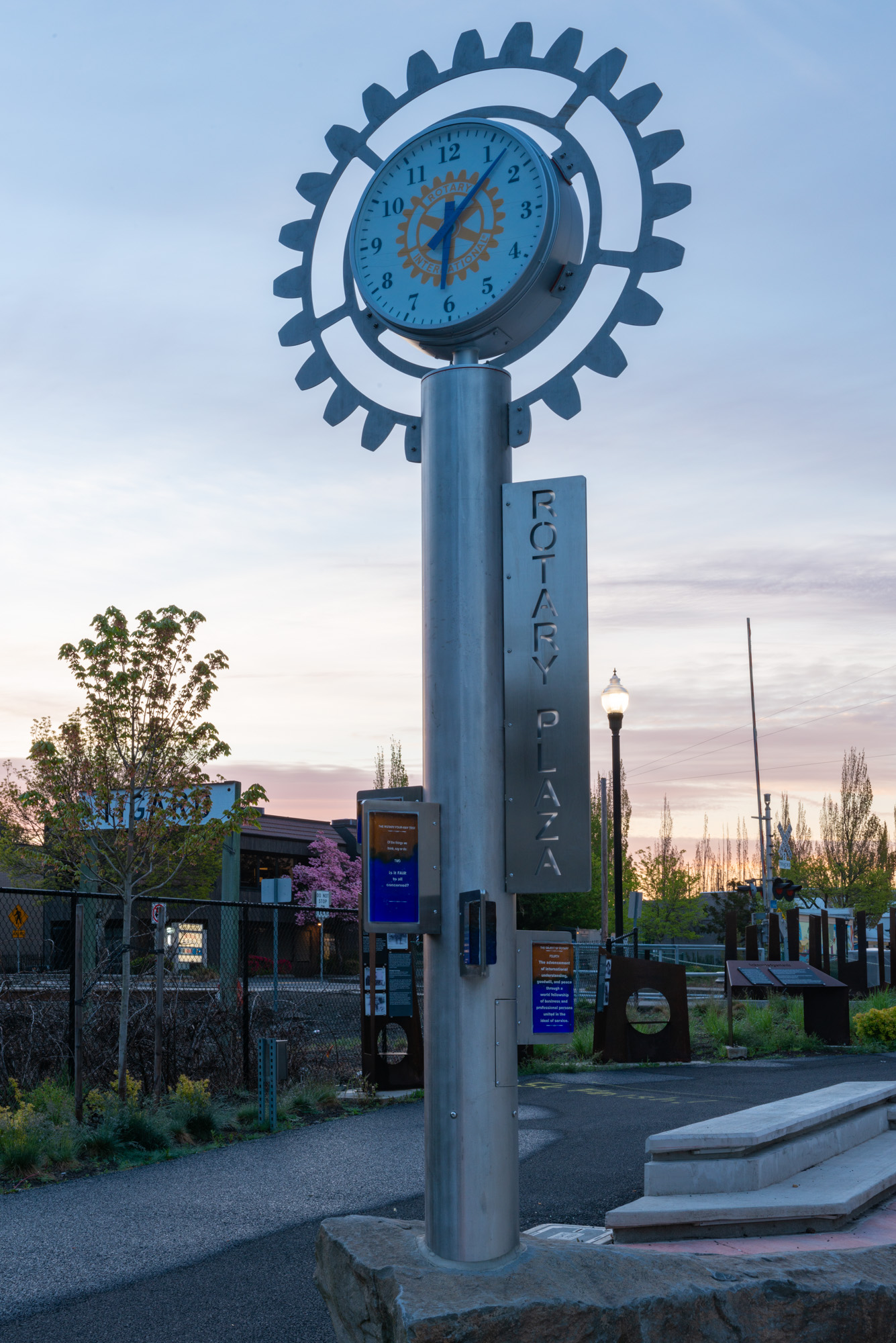 A gear-shaped clock on a metal post at dusk in a downtown public plaza.