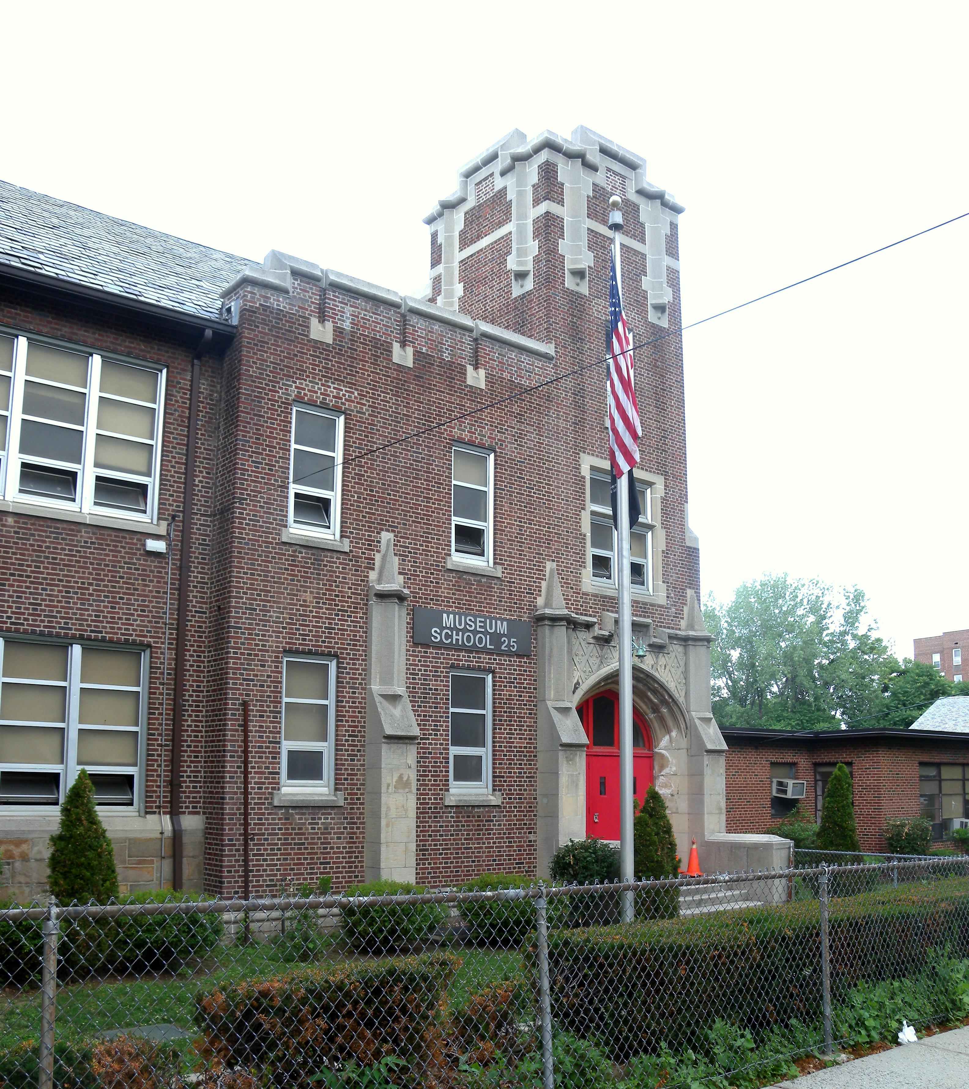 Brick public school building with American flag on flagpole and chain-link fence in foreground.