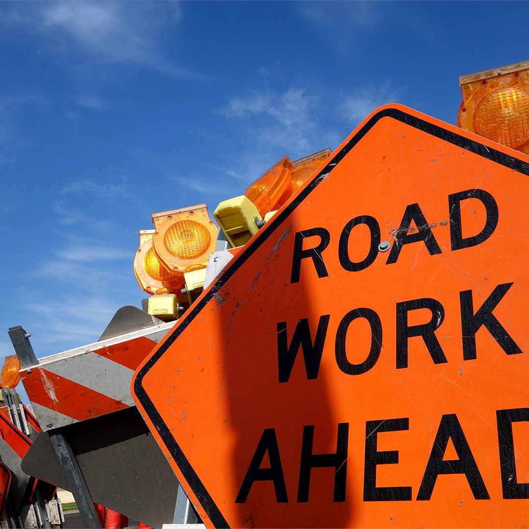 Orange diamond-shaped Road Work Ahead sign with flashing amber barricade lights against a blue sky.