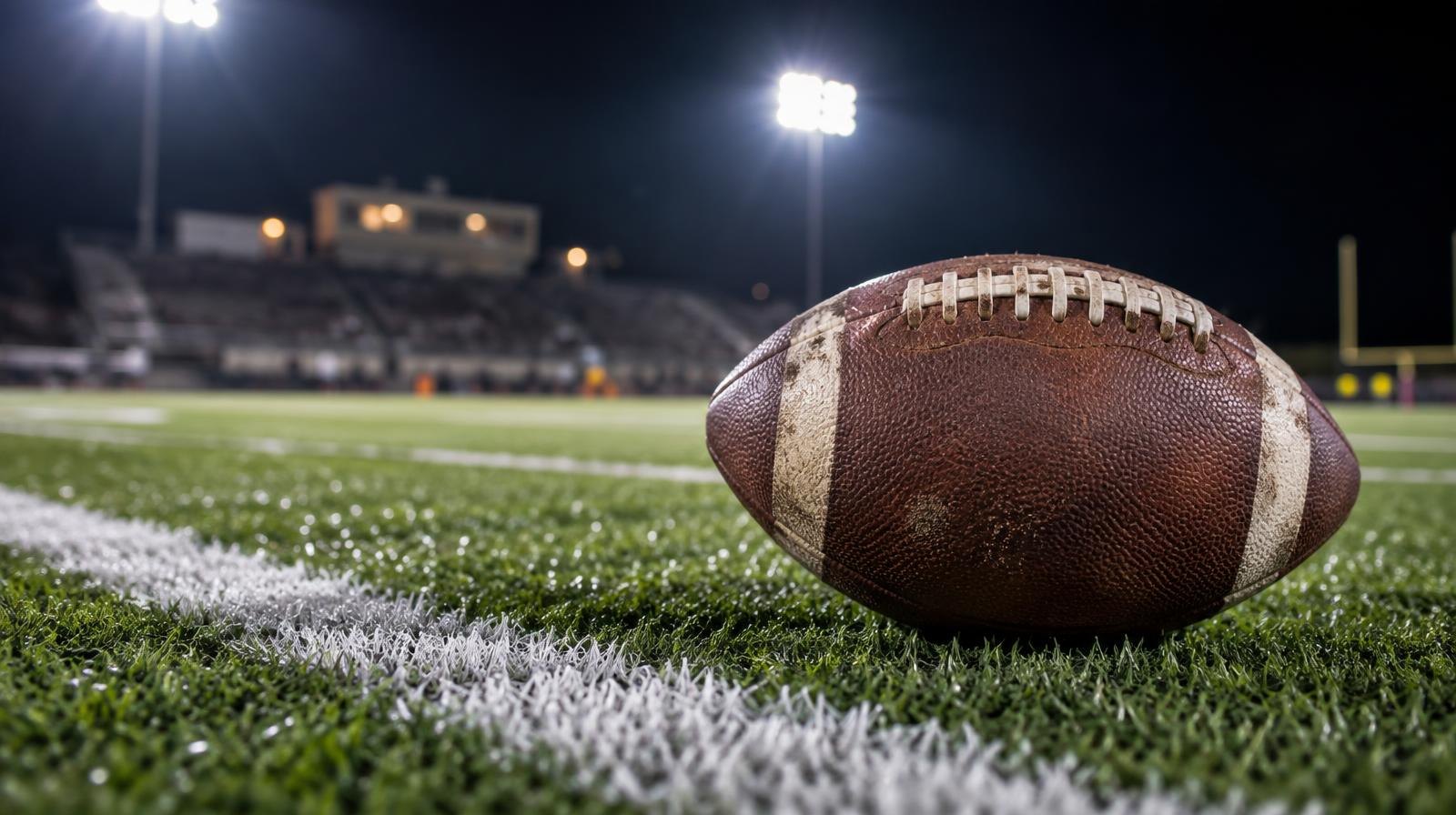 An American football on green turf beside a white yard line under stadium lights.