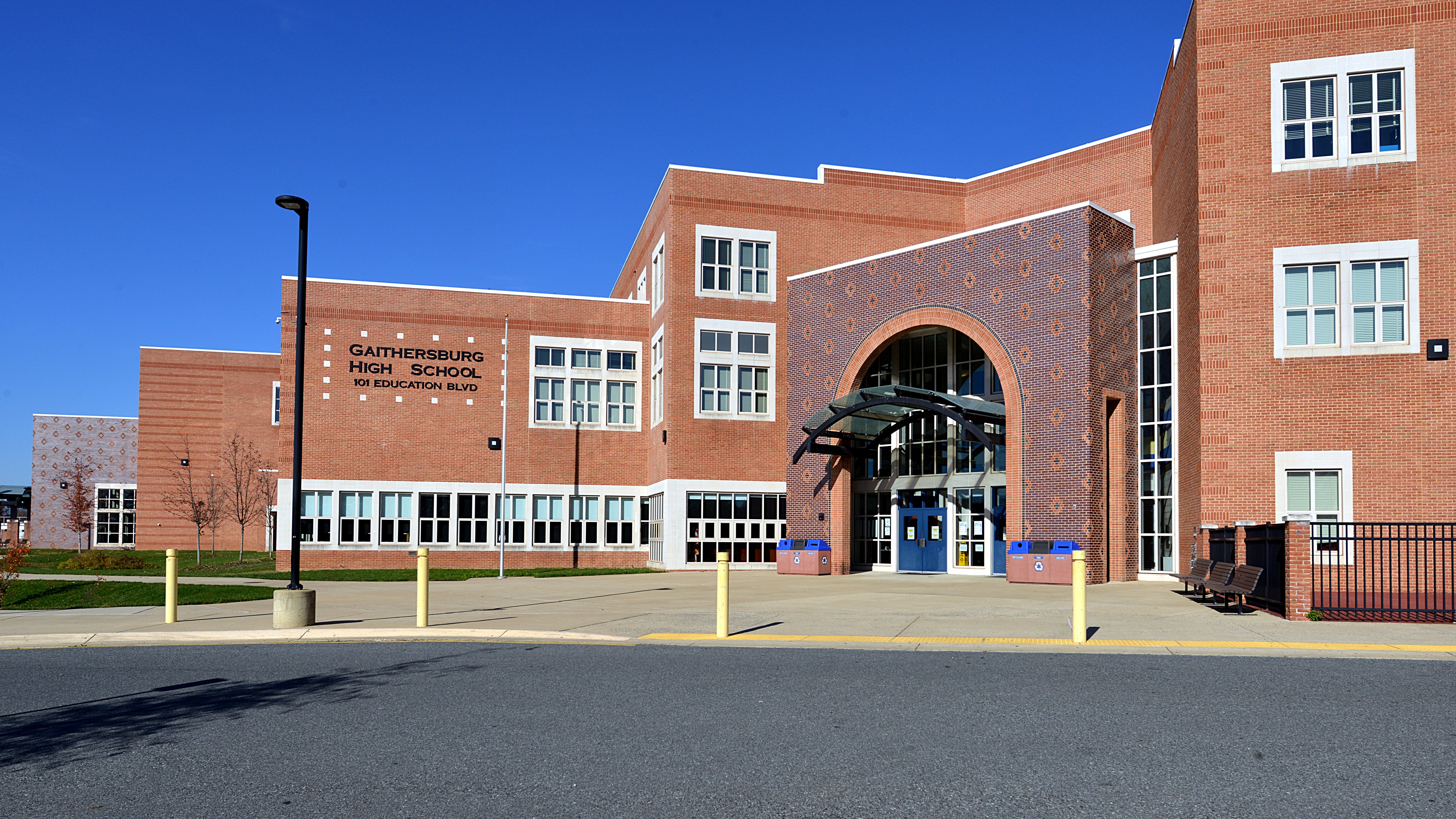 Brick school building with a large arched entrance, blue doors, and yellow bollards under a clear sky.