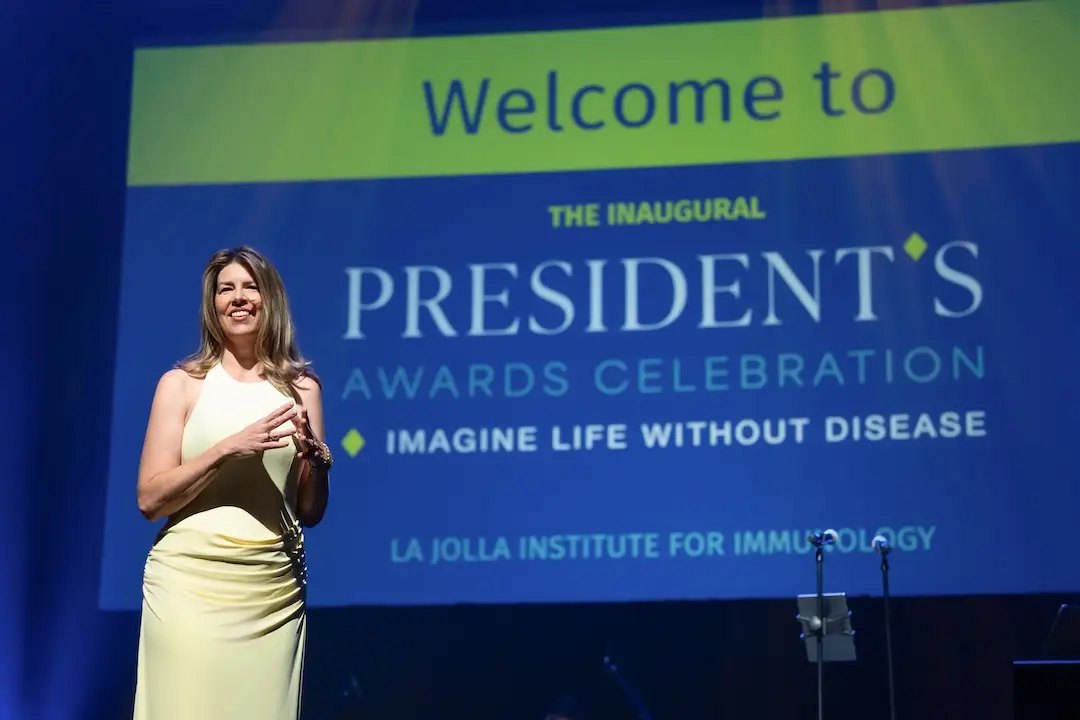 A woman in a yellow dress speaks on a stage in front of a large event screen.