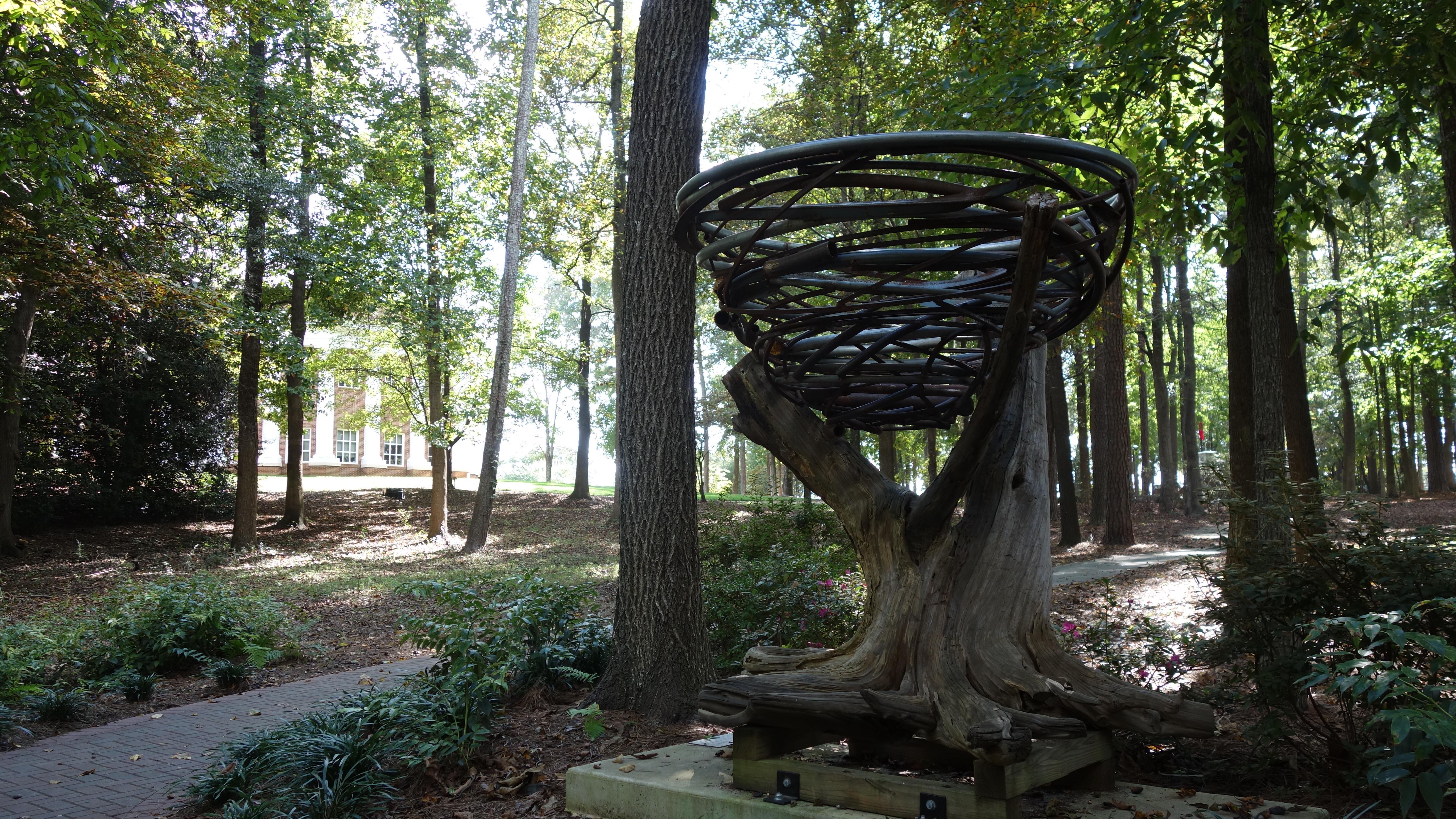 Wooded grounds of a brick civic building with a metal ring sculpture on a tree stump.