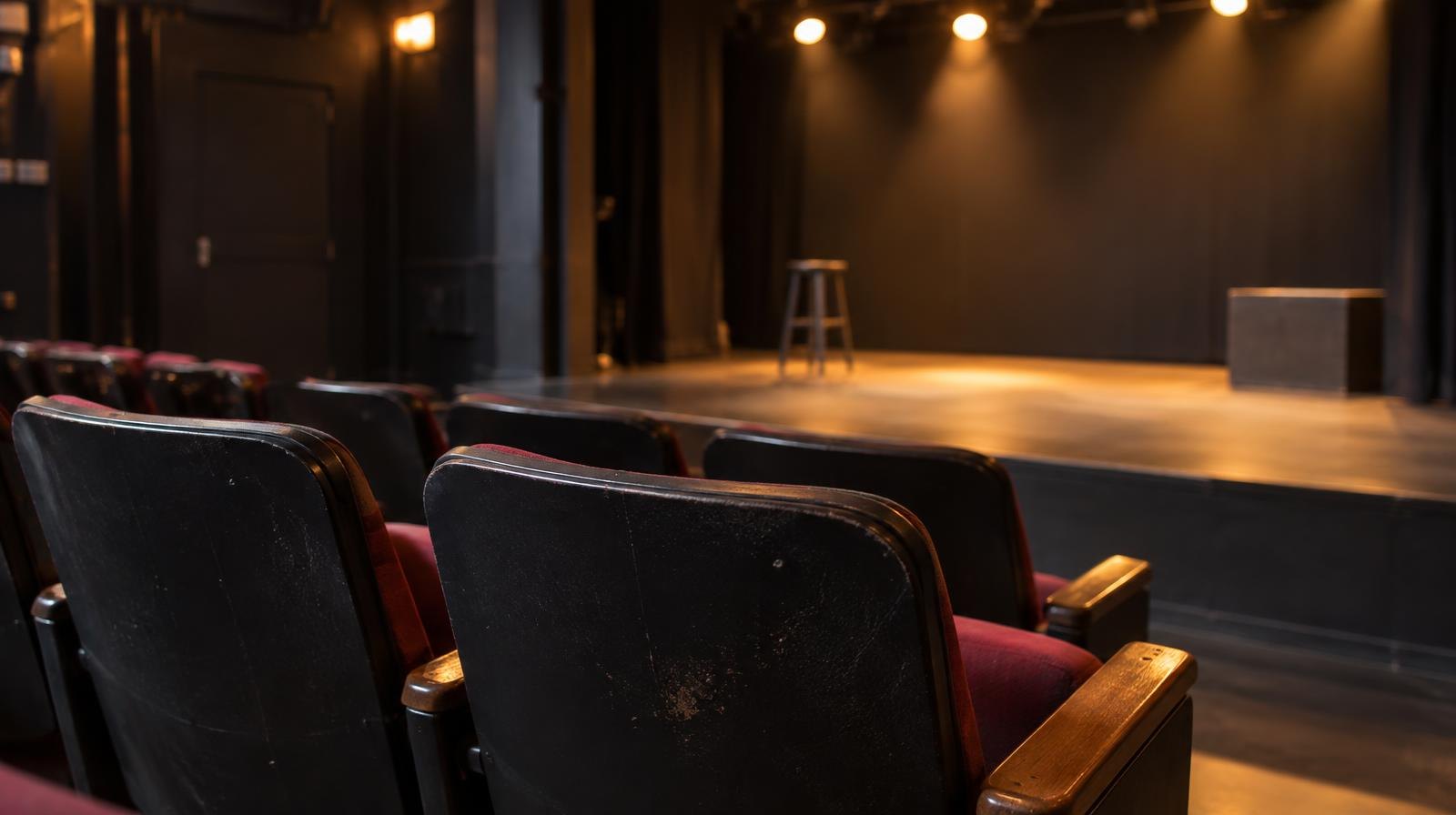 Row of empty seats in a small theater with a warmly lit bare stage in the background.