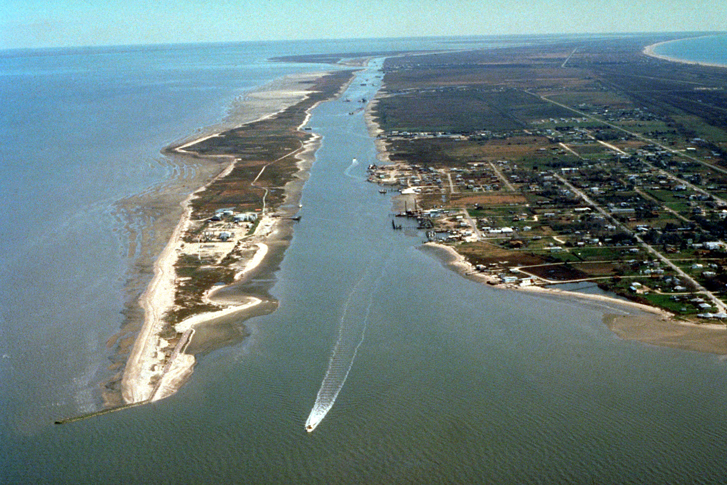 The Gulf Intracoastal Waterway enters Galveston Bay from the southwest end of the Bolivar Peninsula at the town of Port Bolivar, Galveston County, Texas, USA. The community of Port Bolivar lies at right at the end of the peninsula. Galveston Bay is the great body of water at left. A small piece of the Gulf of Mexico can be seen at upper right. The U.S. Army Corps of Engineers maintains and dredges the waterway for commercial barge navigation.