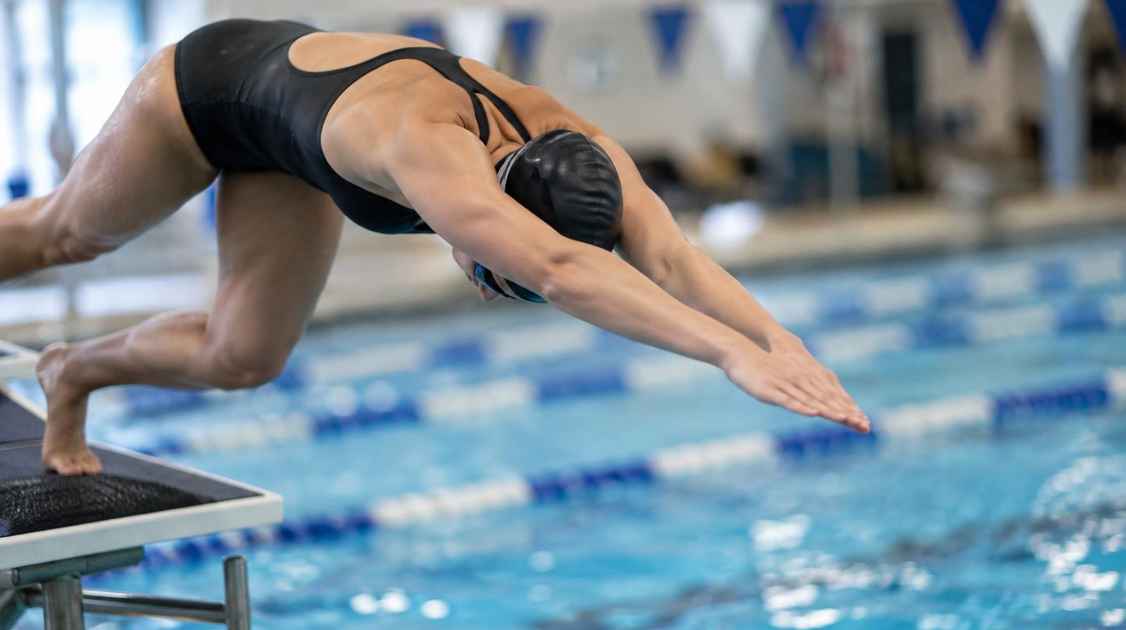 A swimmer in a streamlined dive position launching off a starting block above a blue indoor pool lane.