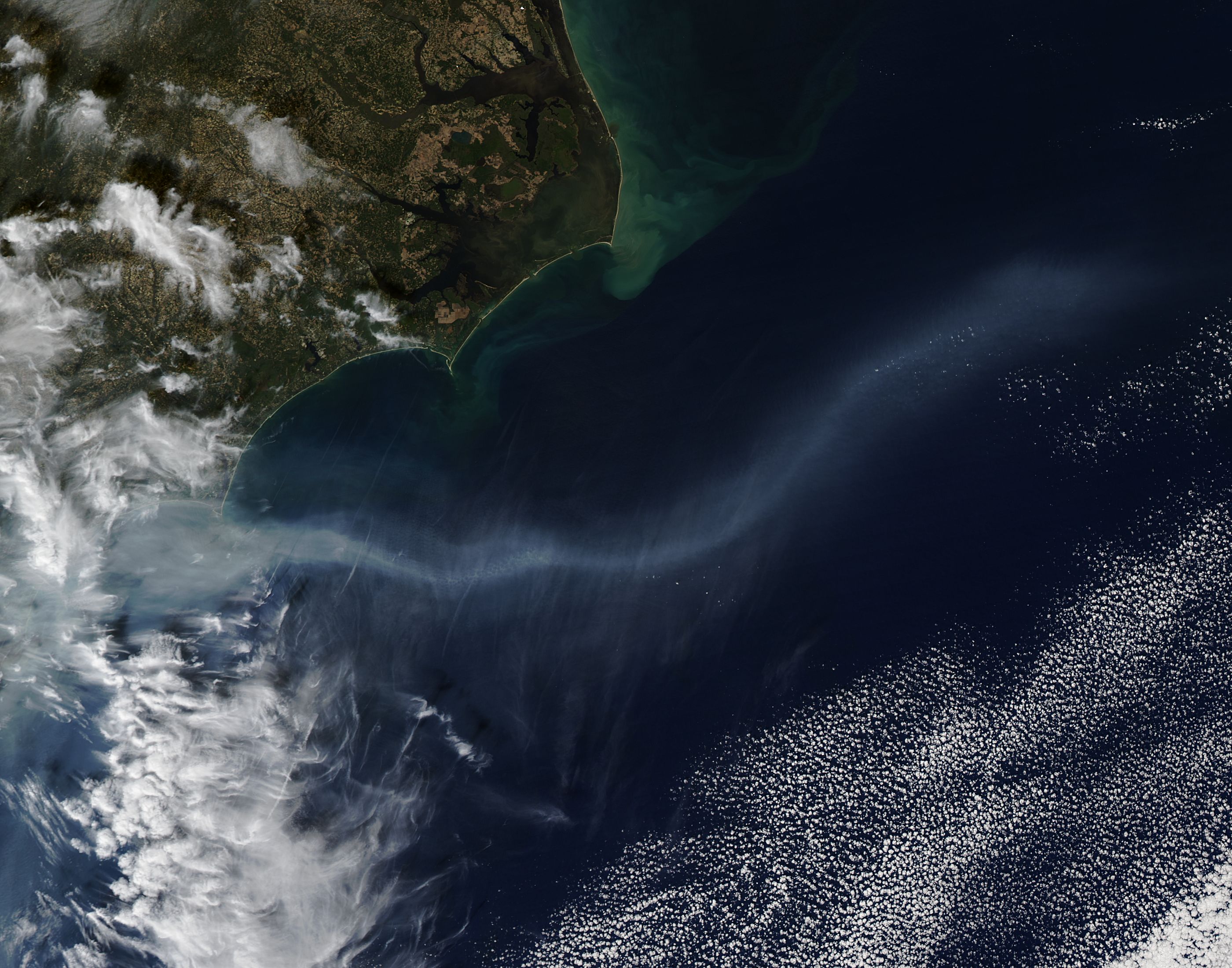 Satellite view of a wide gray smoke plume blowing from a coastline out over dark ocean water.