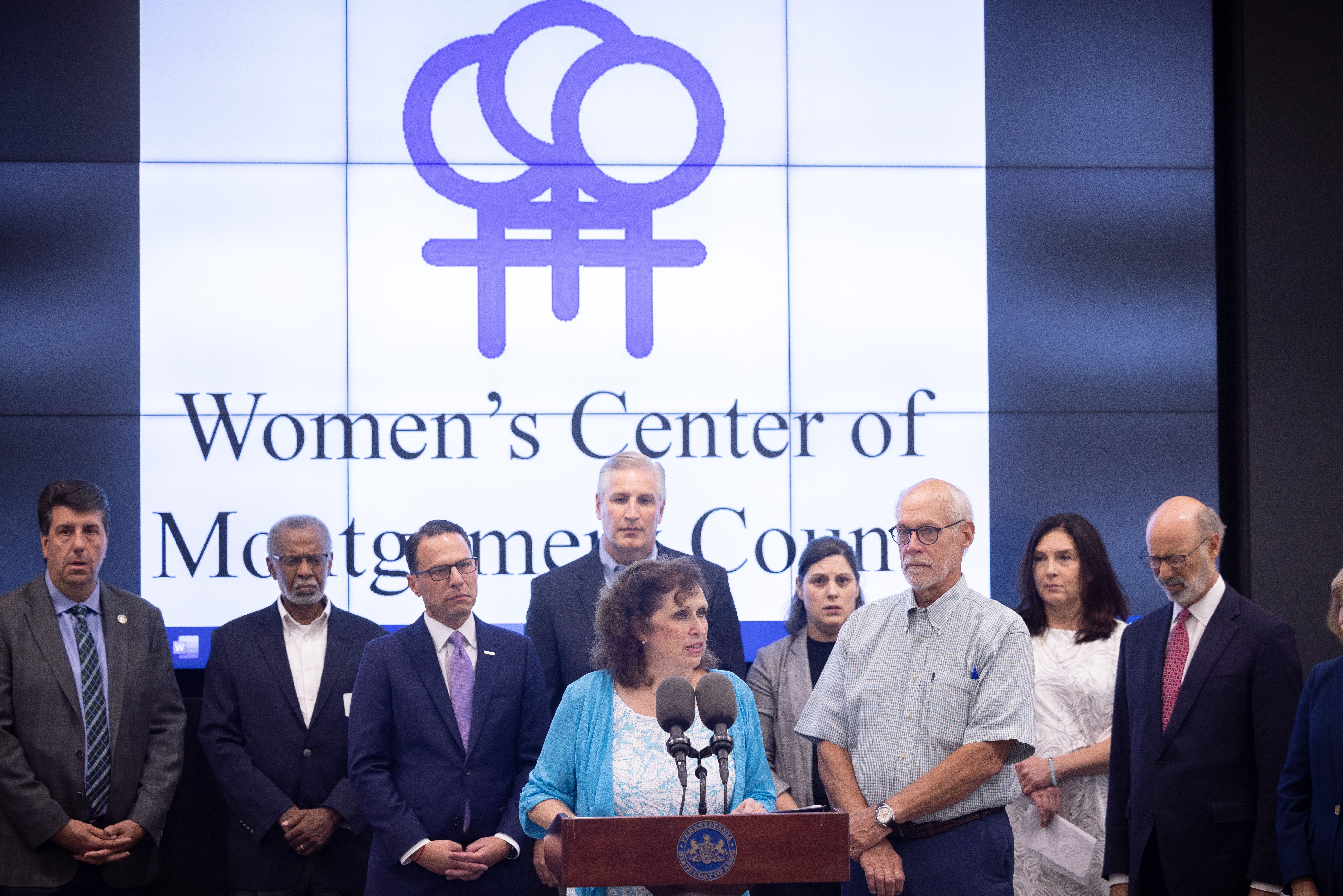 Woman at a podium with an organization logo displayed on a screen behind her.