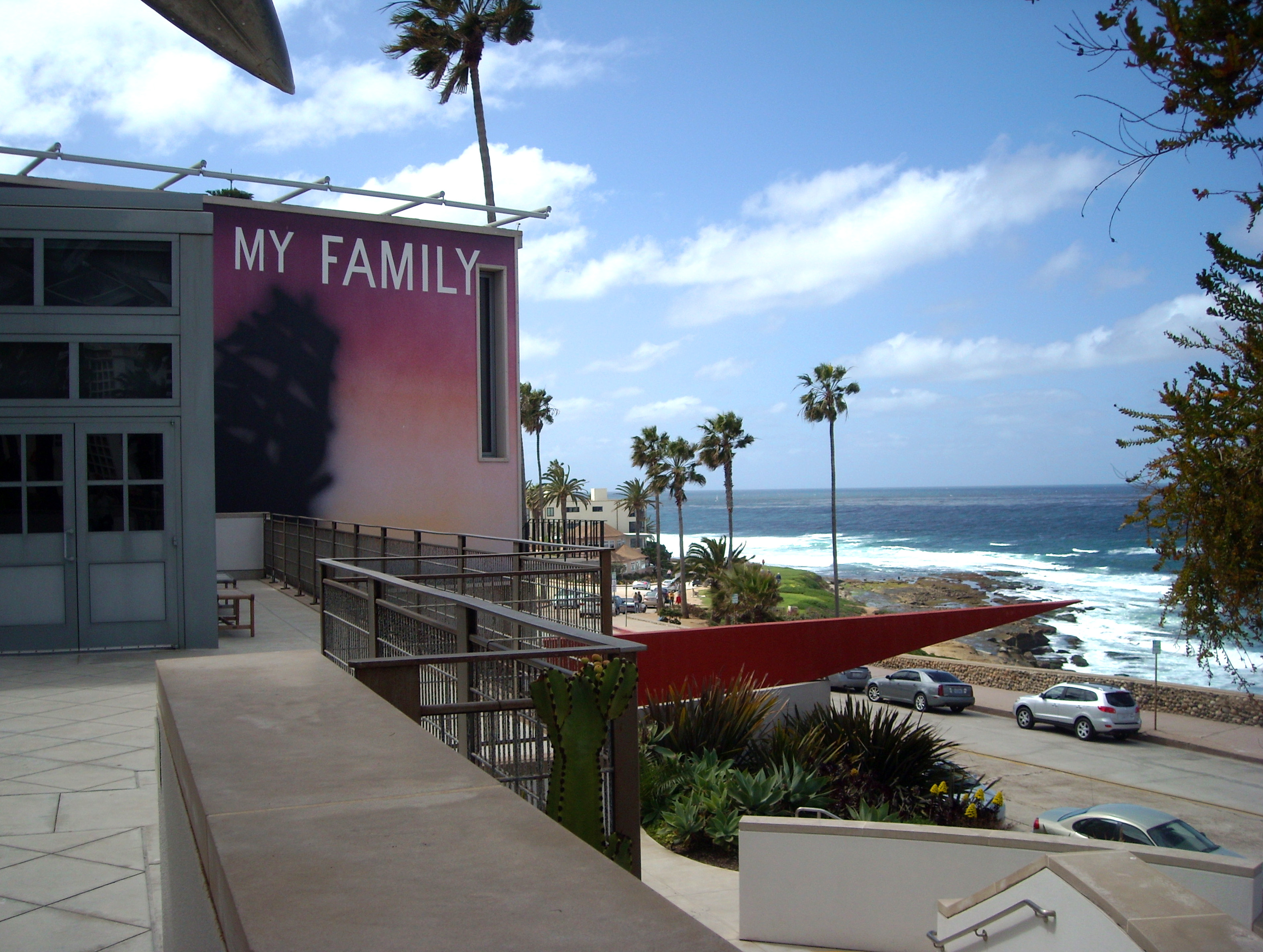 Museum building exterior with a large banner, palm trees, red shade structures, and ocean in the background.