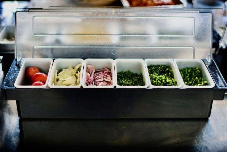 A stainless steel food prep station with six compartments holding tomatoes, sliced vegetables, deli meat, and chopped herbs.