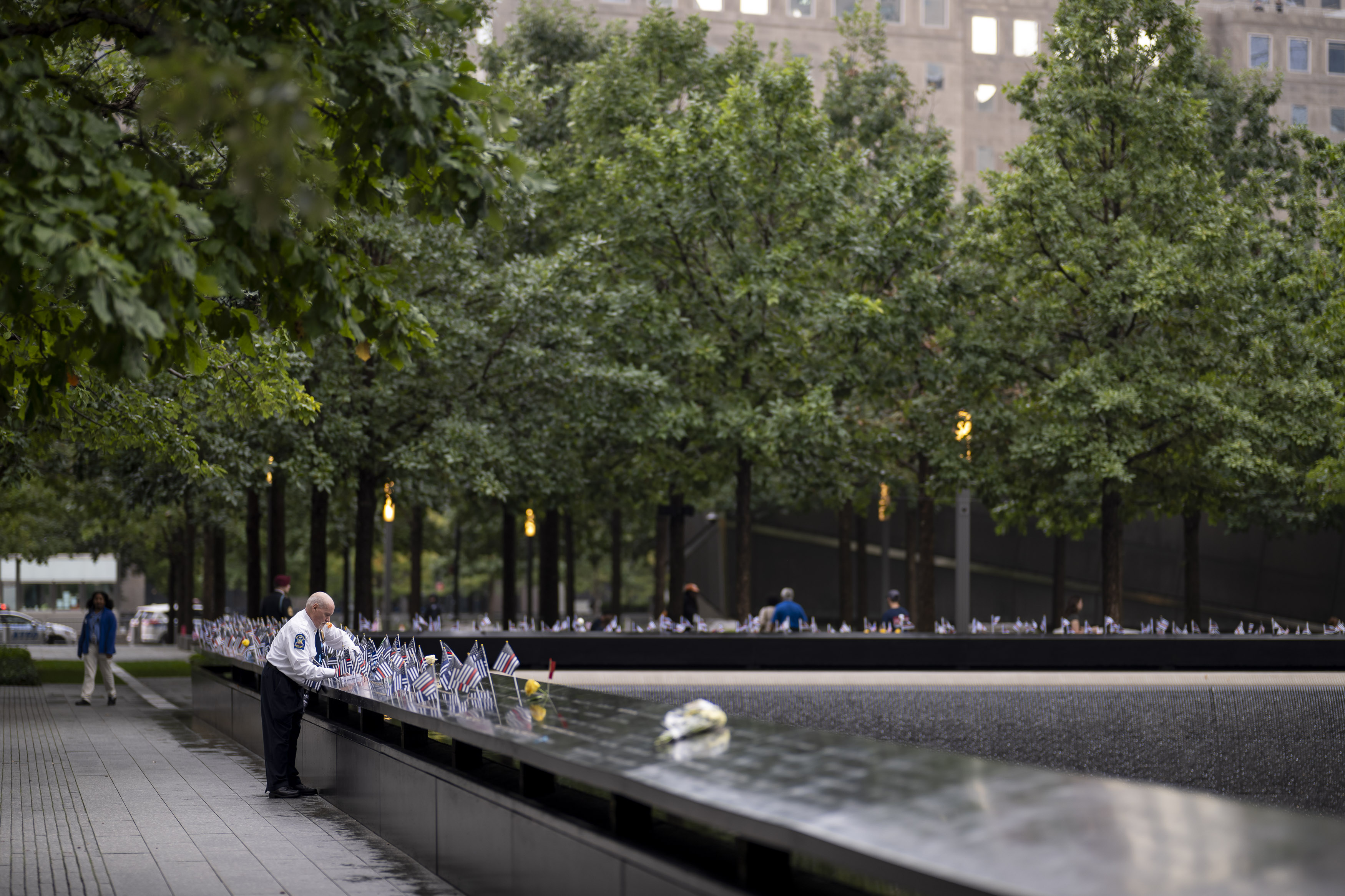 Person standing at a 9/11 Memorial reflecting pool lined with small American flags and flowers.