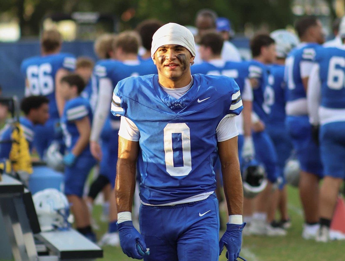 Football player in blue jersey number 0 and white skull cap walking along the sideline with teammates behind him.