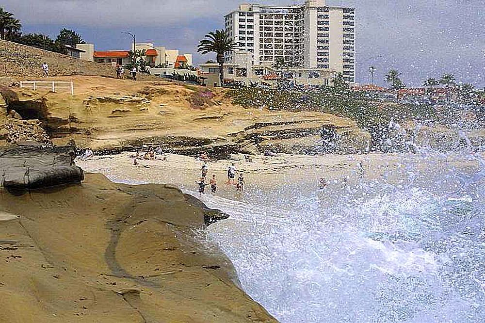 Large ocean waves crash against rocky sandstone cliffs at a California beach.