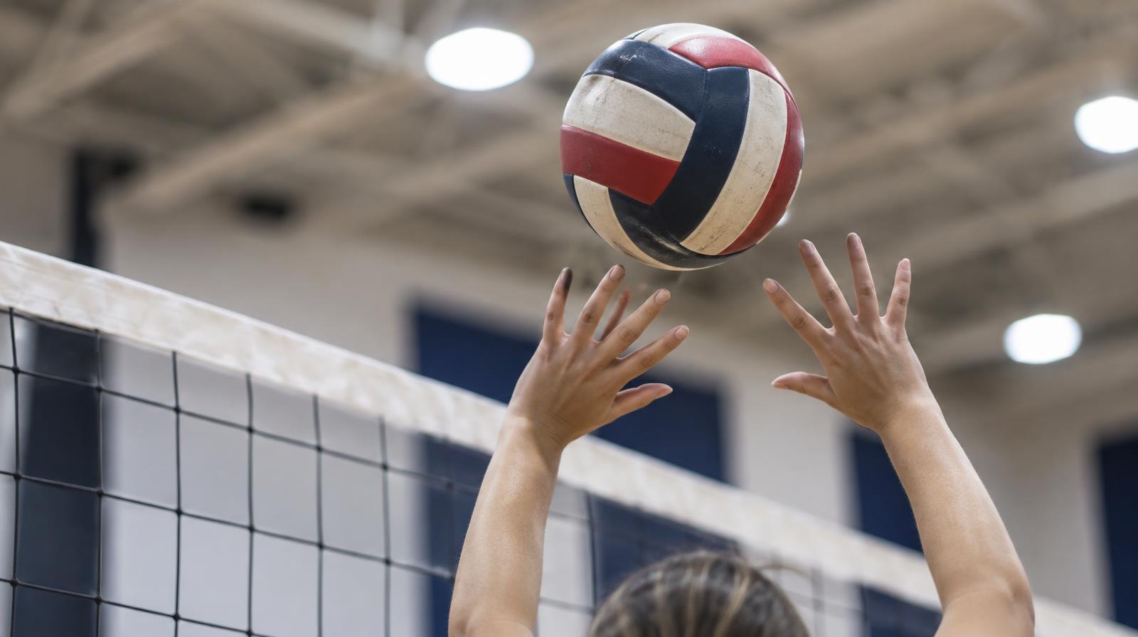 Hands setting a volleyball above the net in a brightly lit gymnasium.