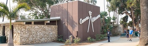 Brown mid-century modern Camera Obscura building with stone base, palm trees, and pedestrians nearby.