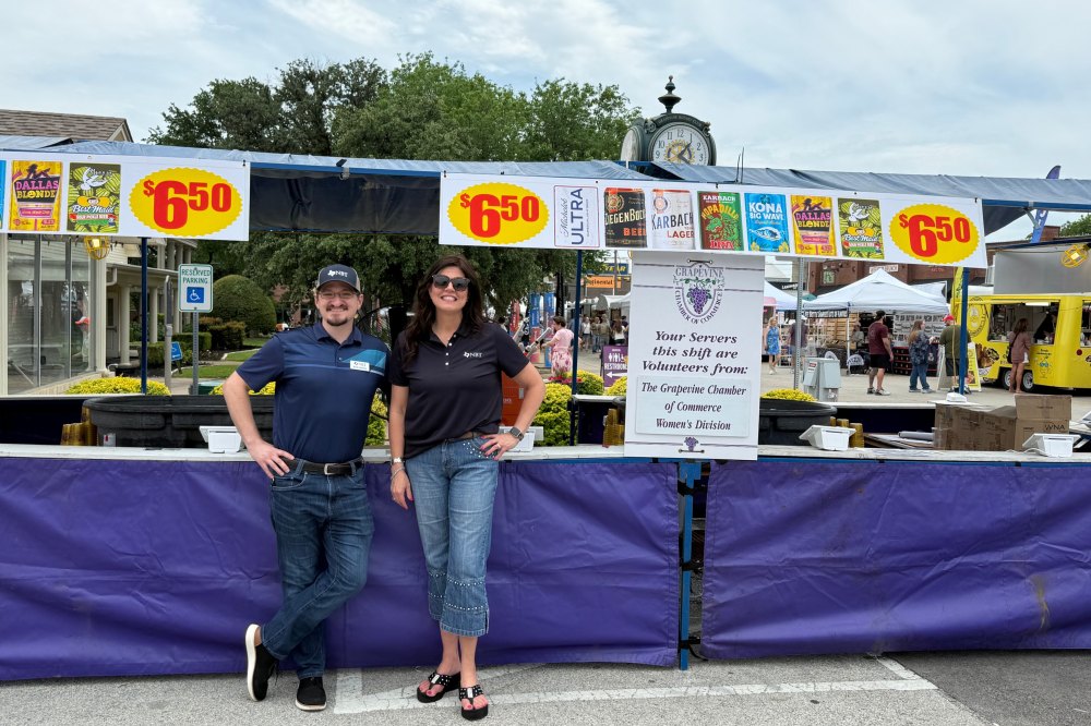 Two people in matching dark polo shirts stand at an outdoor vendor booth with community event signage.