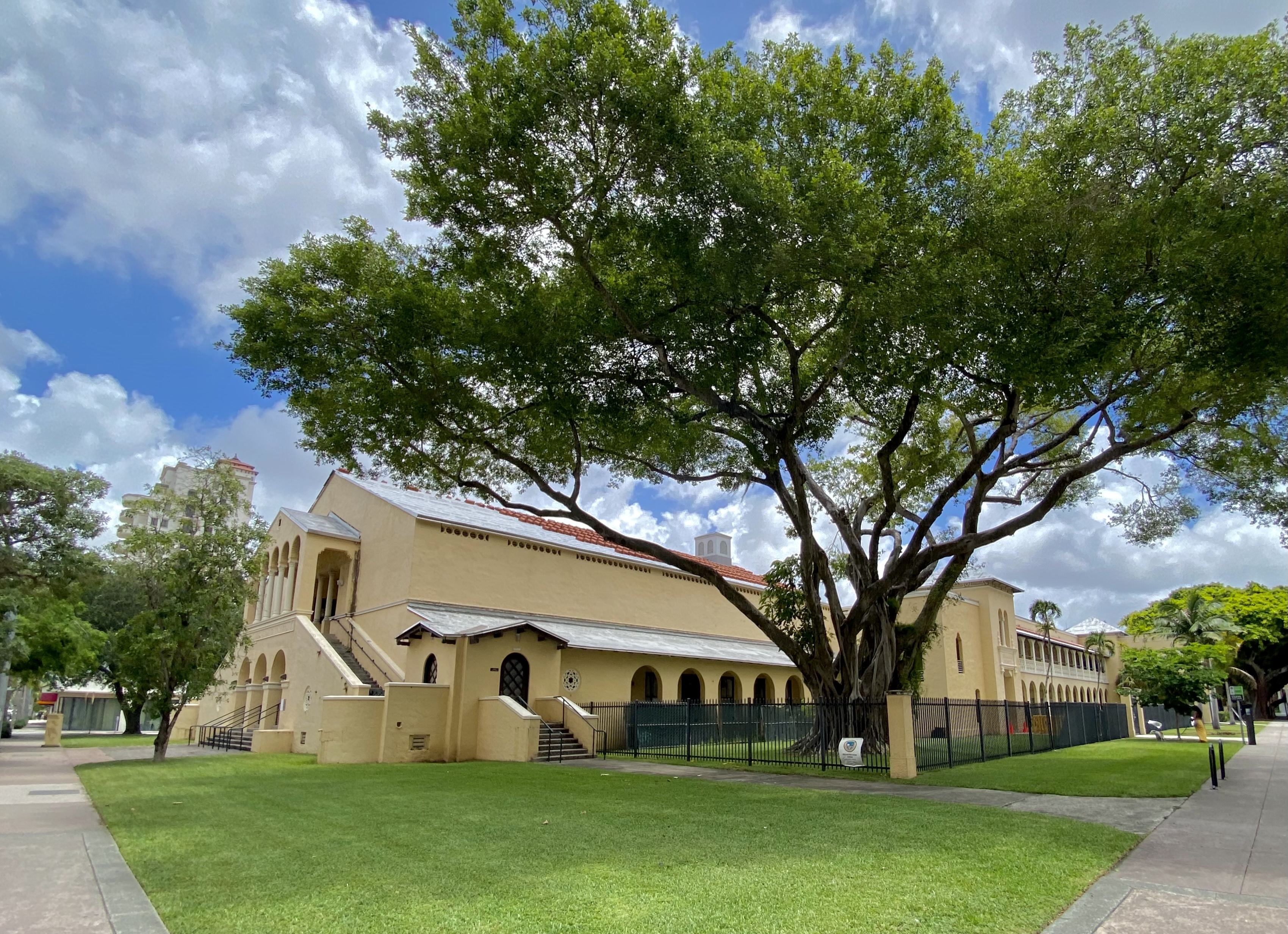 Yellow stucco school building with arched windows and a large banyan tree on a green lawn.