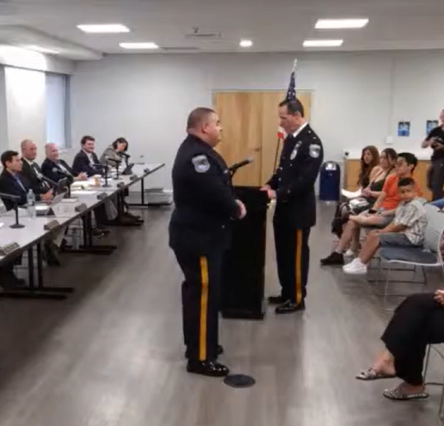 Two police officers in dress uniforms facing each other during a formal ceremony in a meeting room with an American flag.