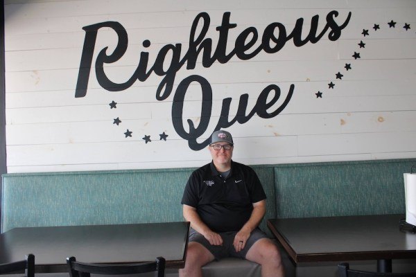 Man in black polo and cap seated in a booth beneath a large Righteous Que wall sign.