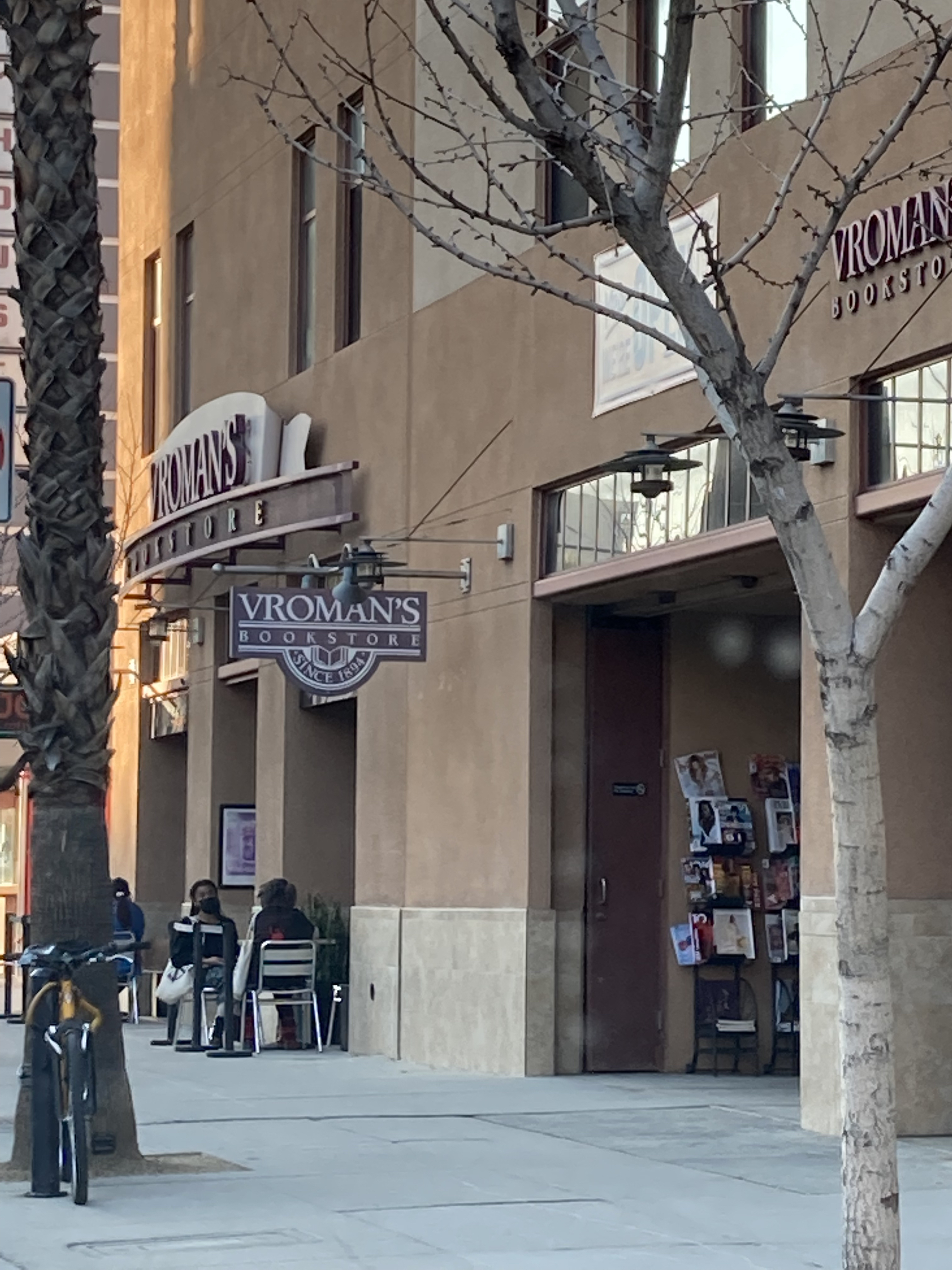 Exterior of a bookstore with large signage, a book display rack, and people at outdoor sidewalk tables.