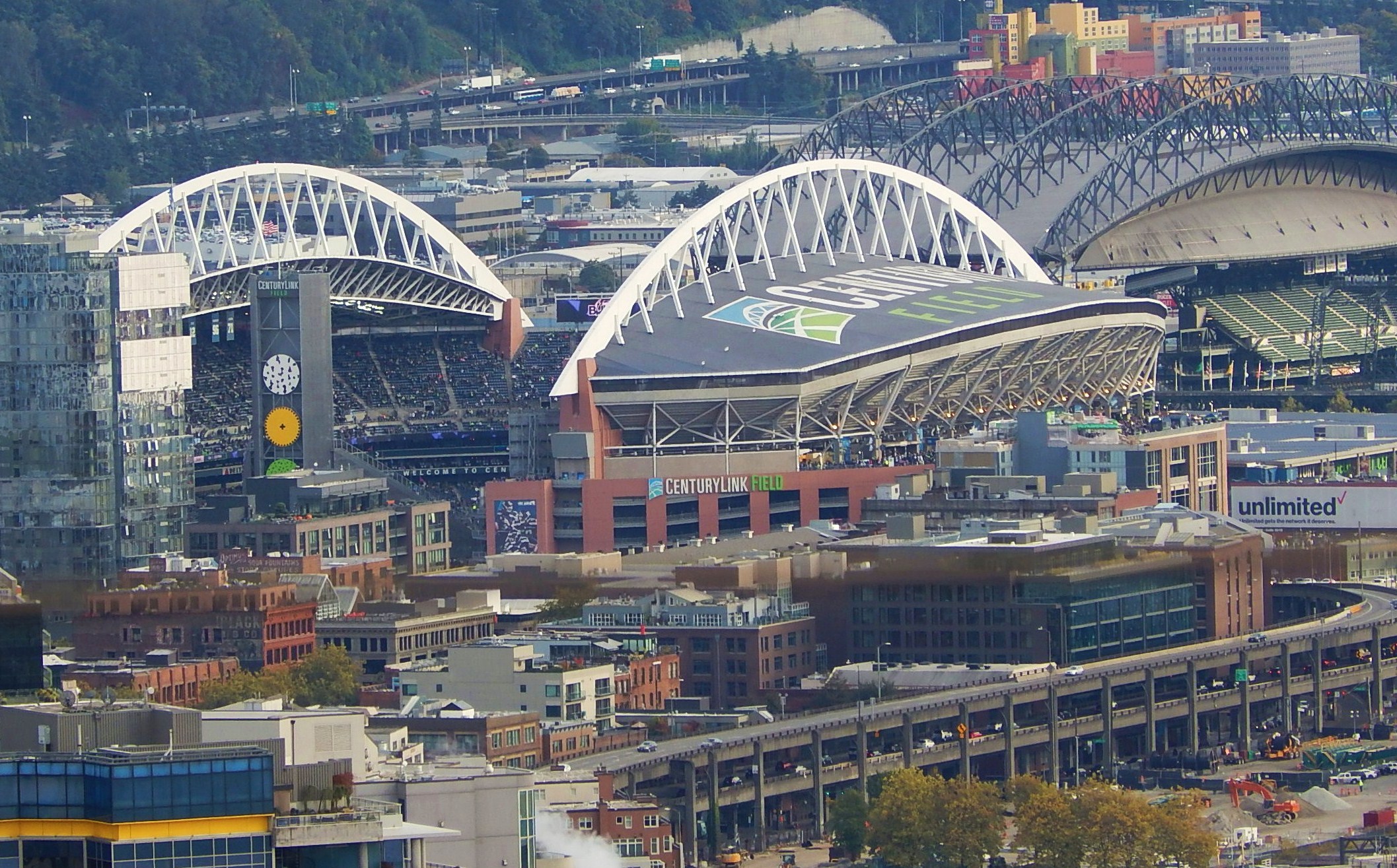 Aerial view of a football stadium with arched roof and adjacent domed stadium in a city skyline.