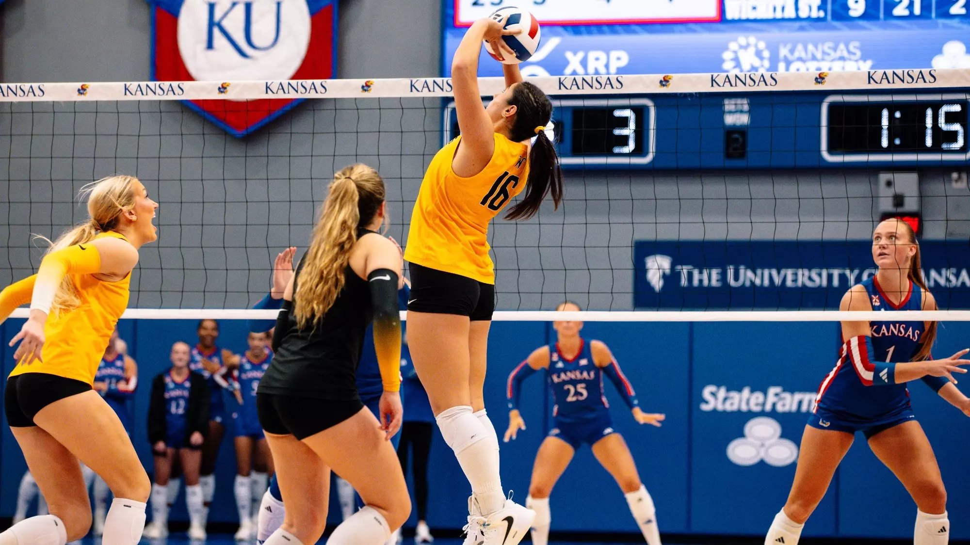 A volleyball player in a gold uniform jumps to attack the ball at the net.