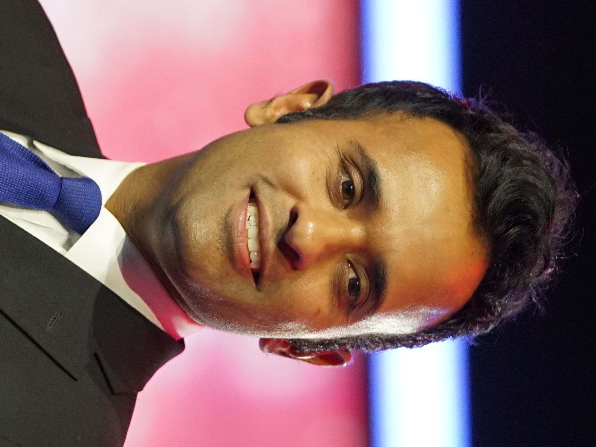 Smiling man in a dark suit and blue tie against a blurred pink-red background.