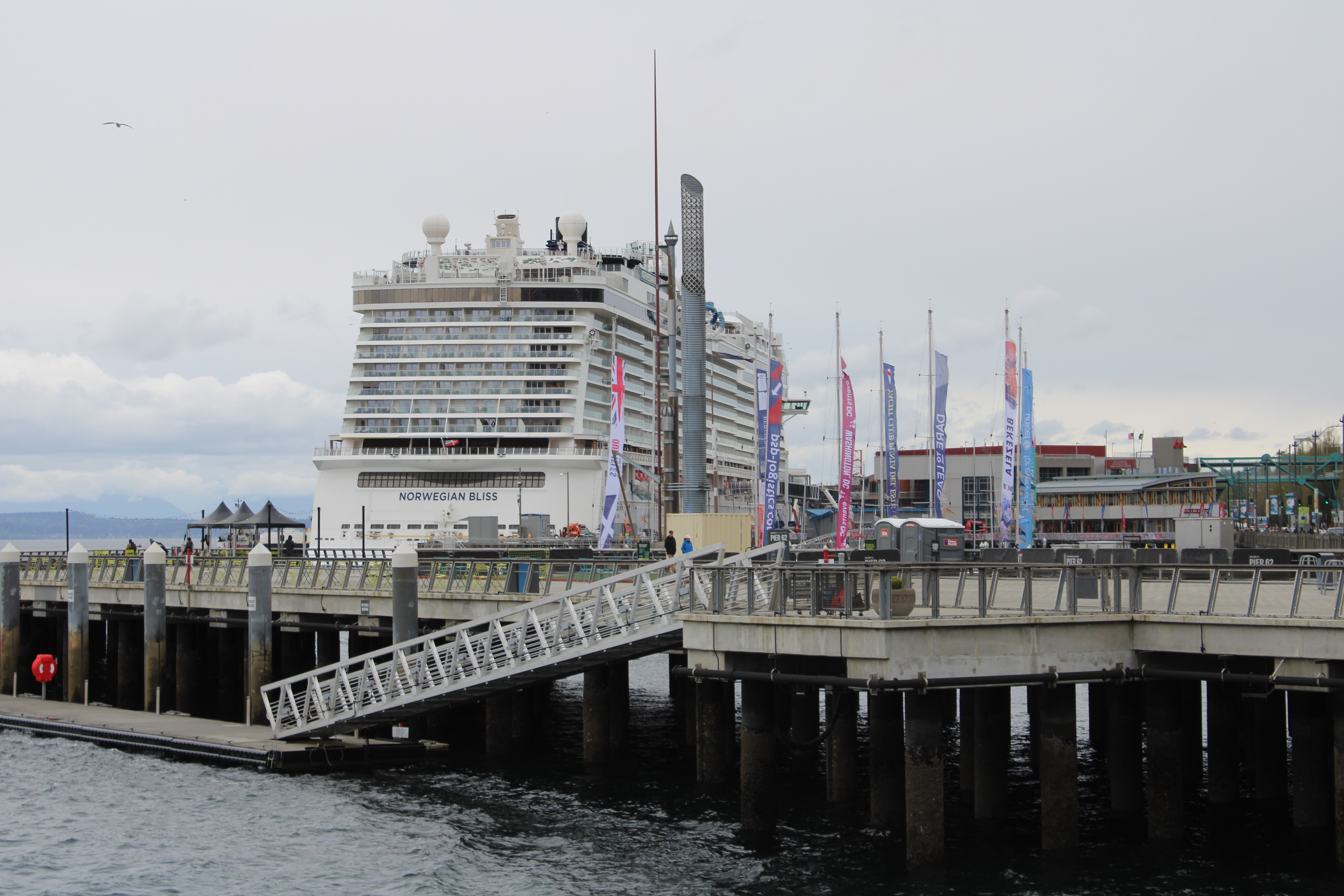 Large cruise ship docked at a pier with colorful flags and a pedestrian walkway under overcast skies