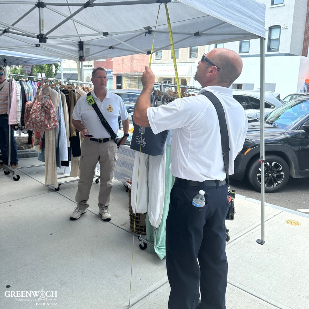 Two uniformed officials inspecting a pop-up vendor tent on a downtown sidewalk with clothing racks nearby.
