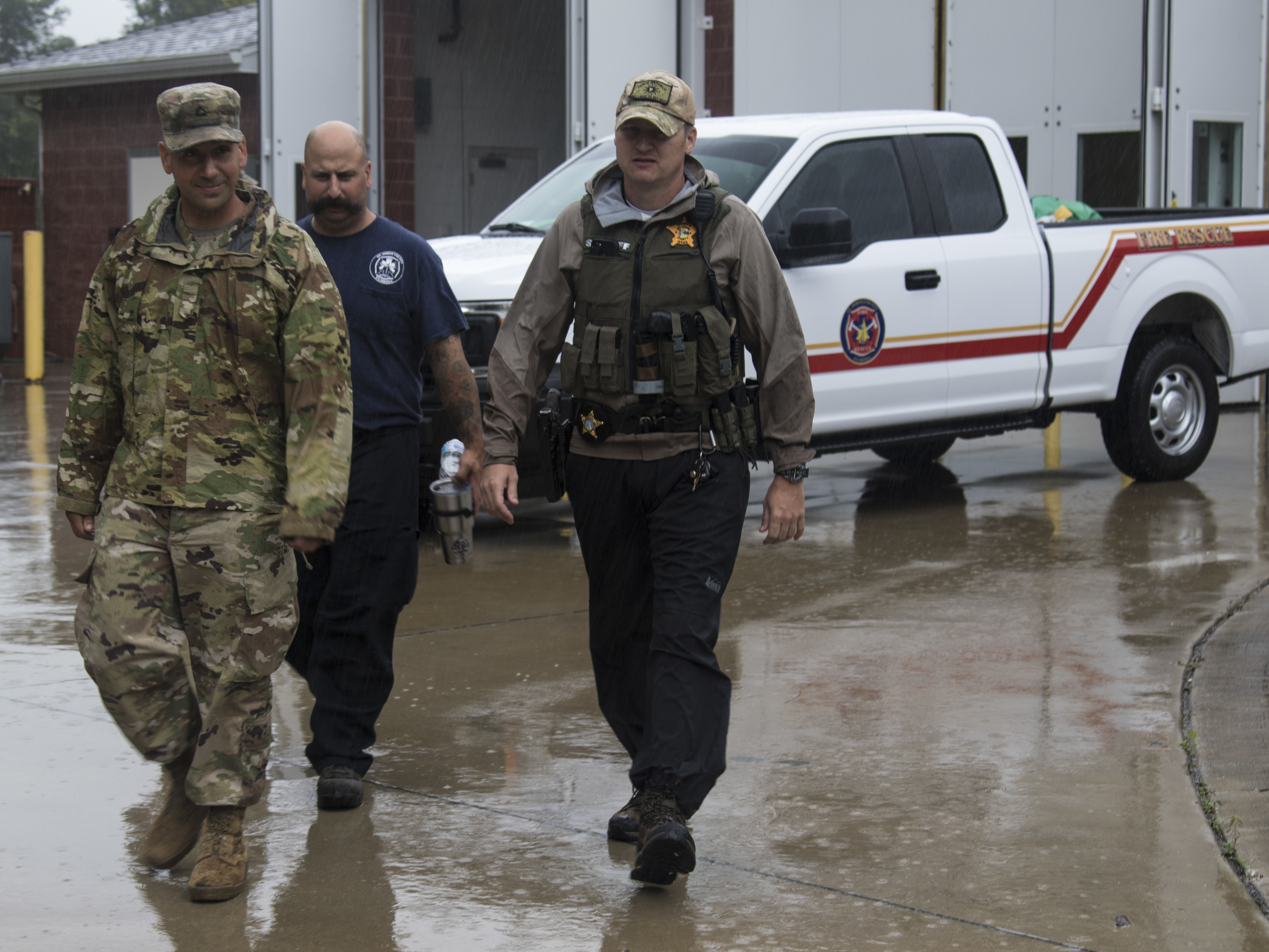 Three uniformed personnel walking on wet pavement near a white fire rescue truck.