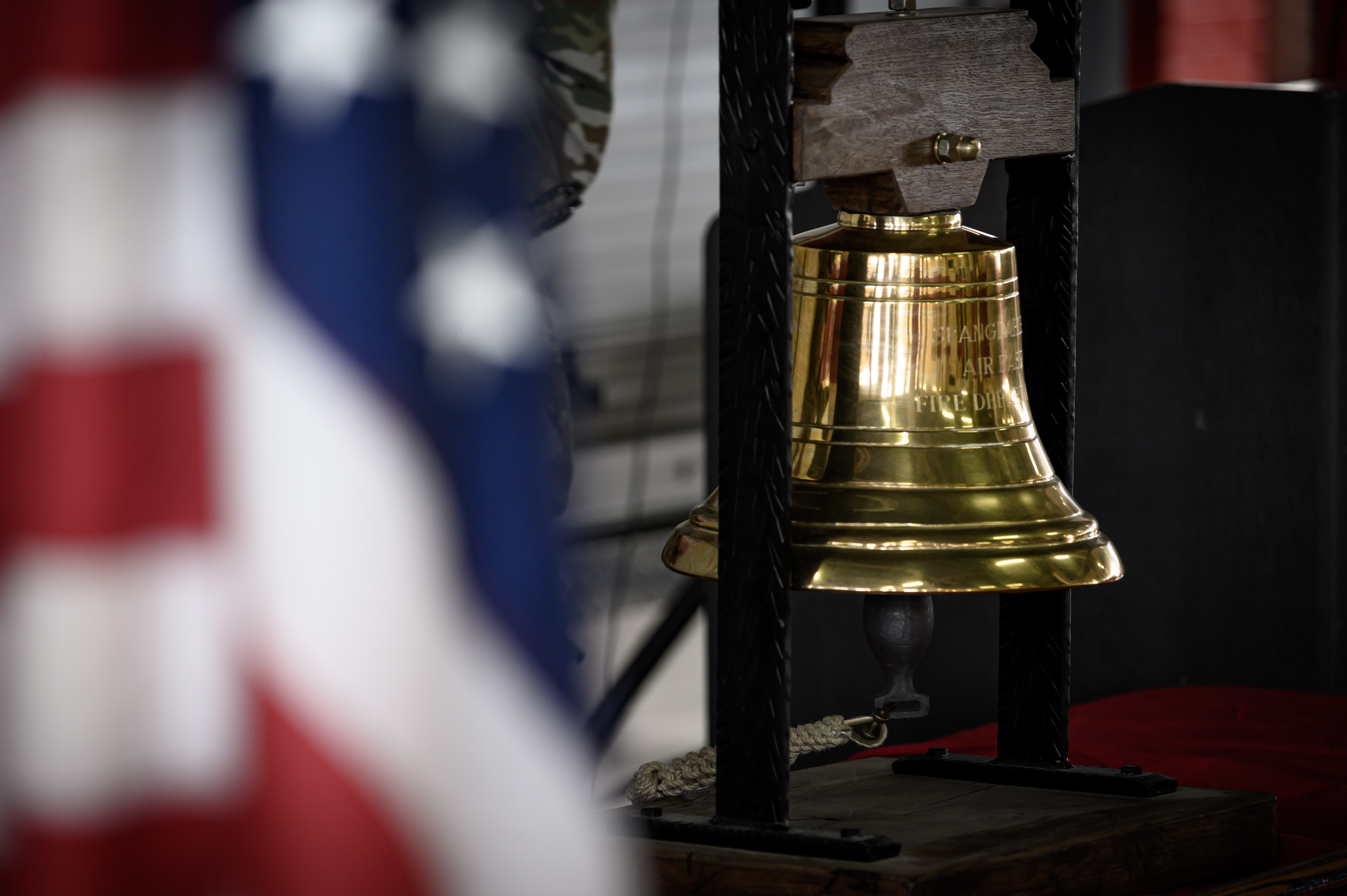A brass ceremonial bell on a stand beside a blurred American flag.