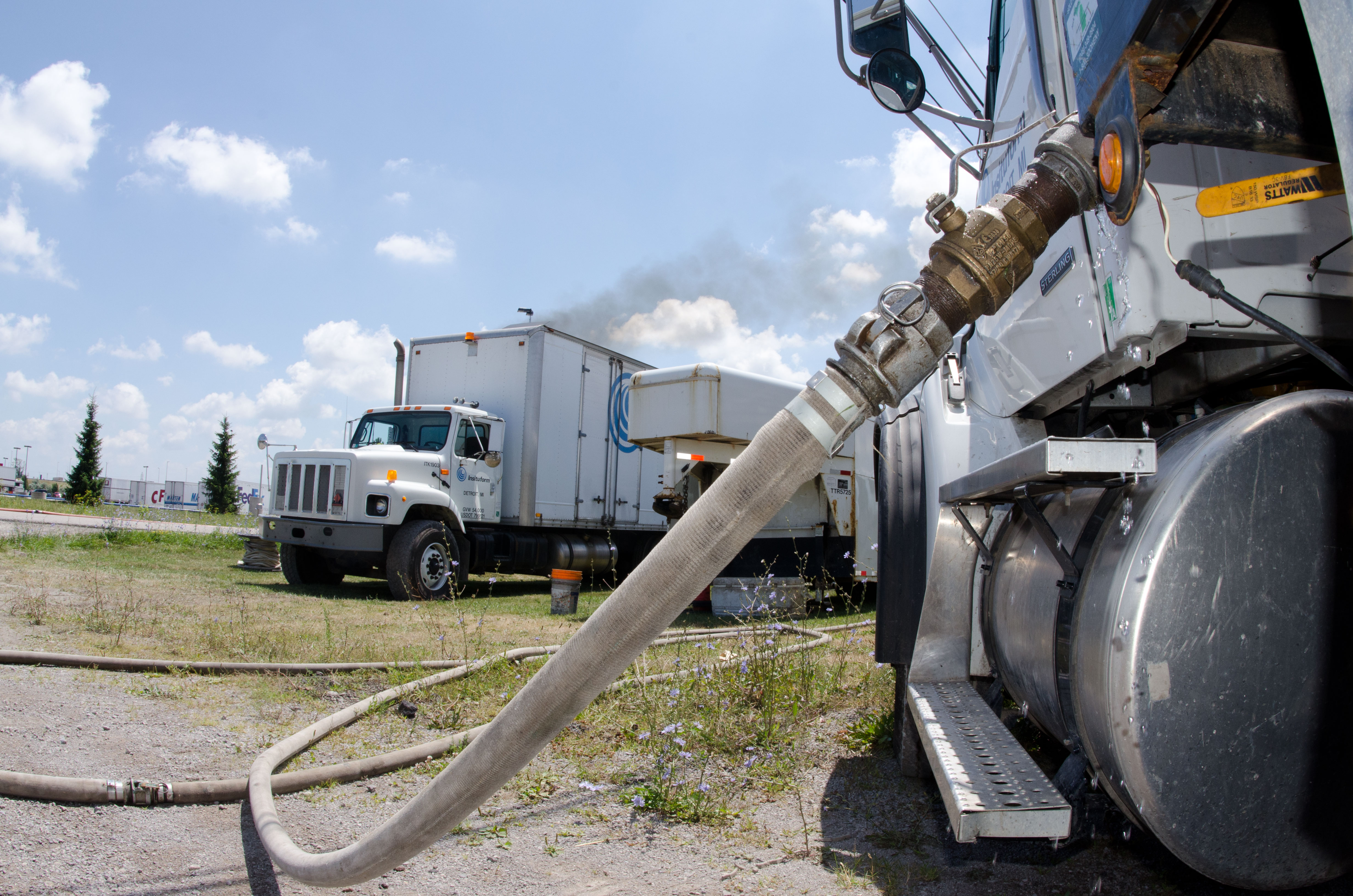 Large hoses connect a tanker truck to a boiler truck at a sewer work site.