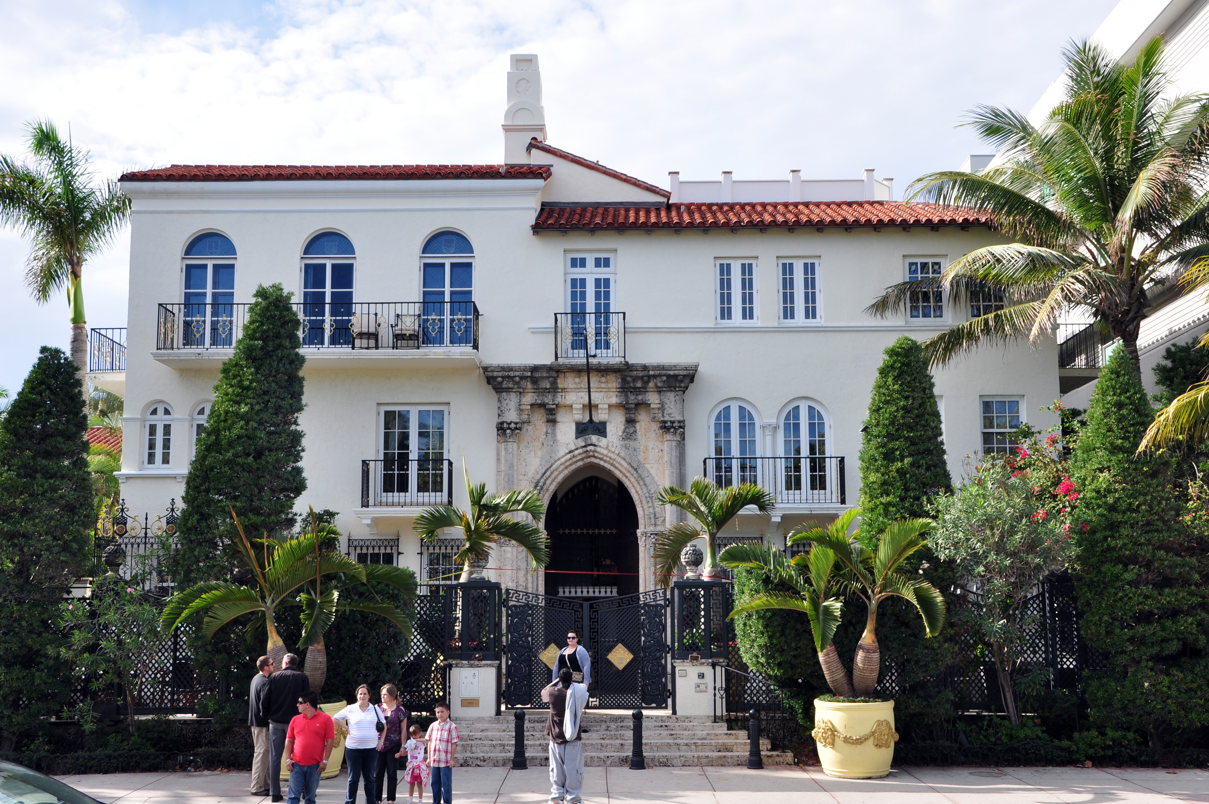 White Mediterranean mansion with red tile roof, iron gates, and palm trees on a sunny street.