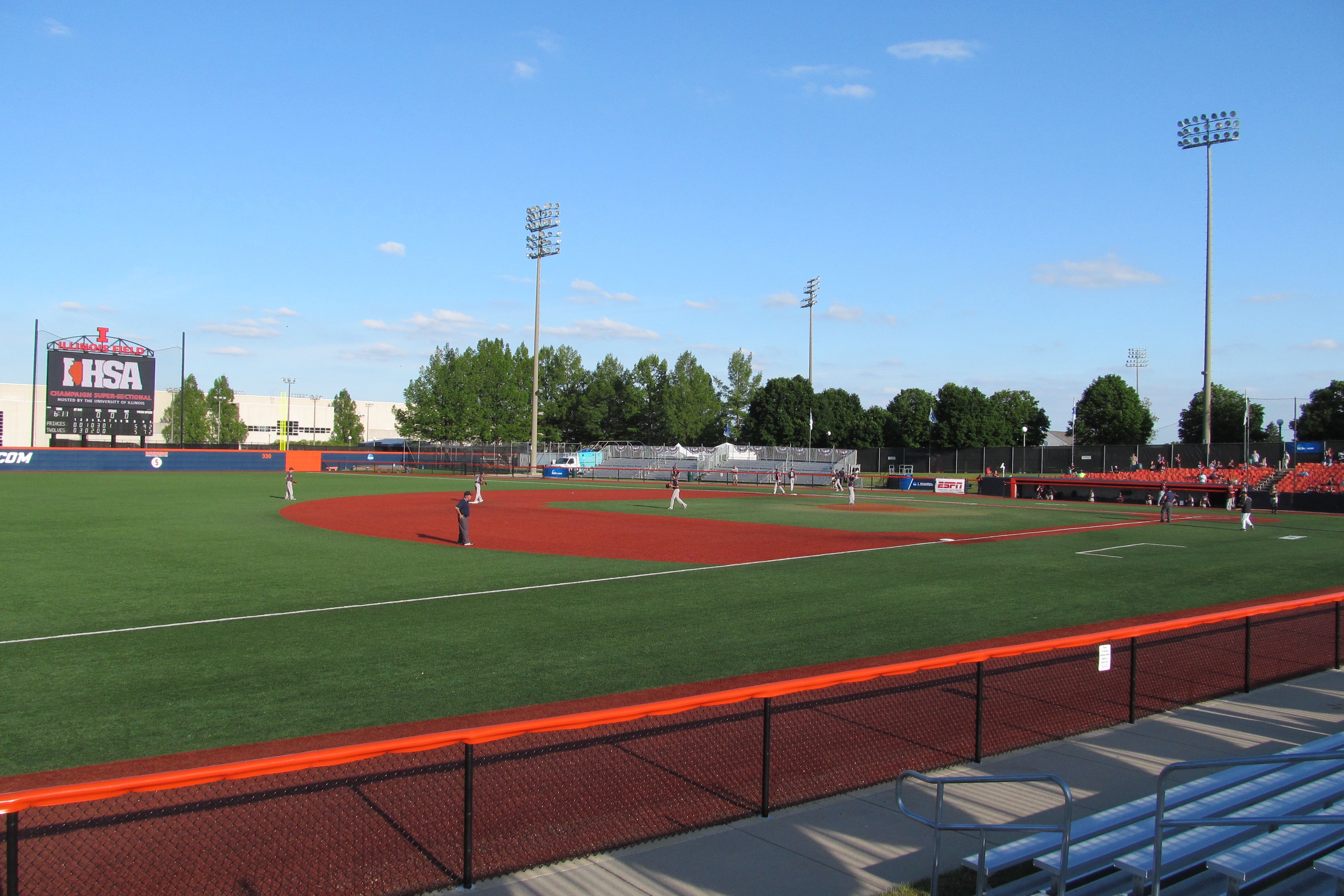 A baseball game in progress on a turf field with blue bleachers and IHSA logo on the outfield wall.