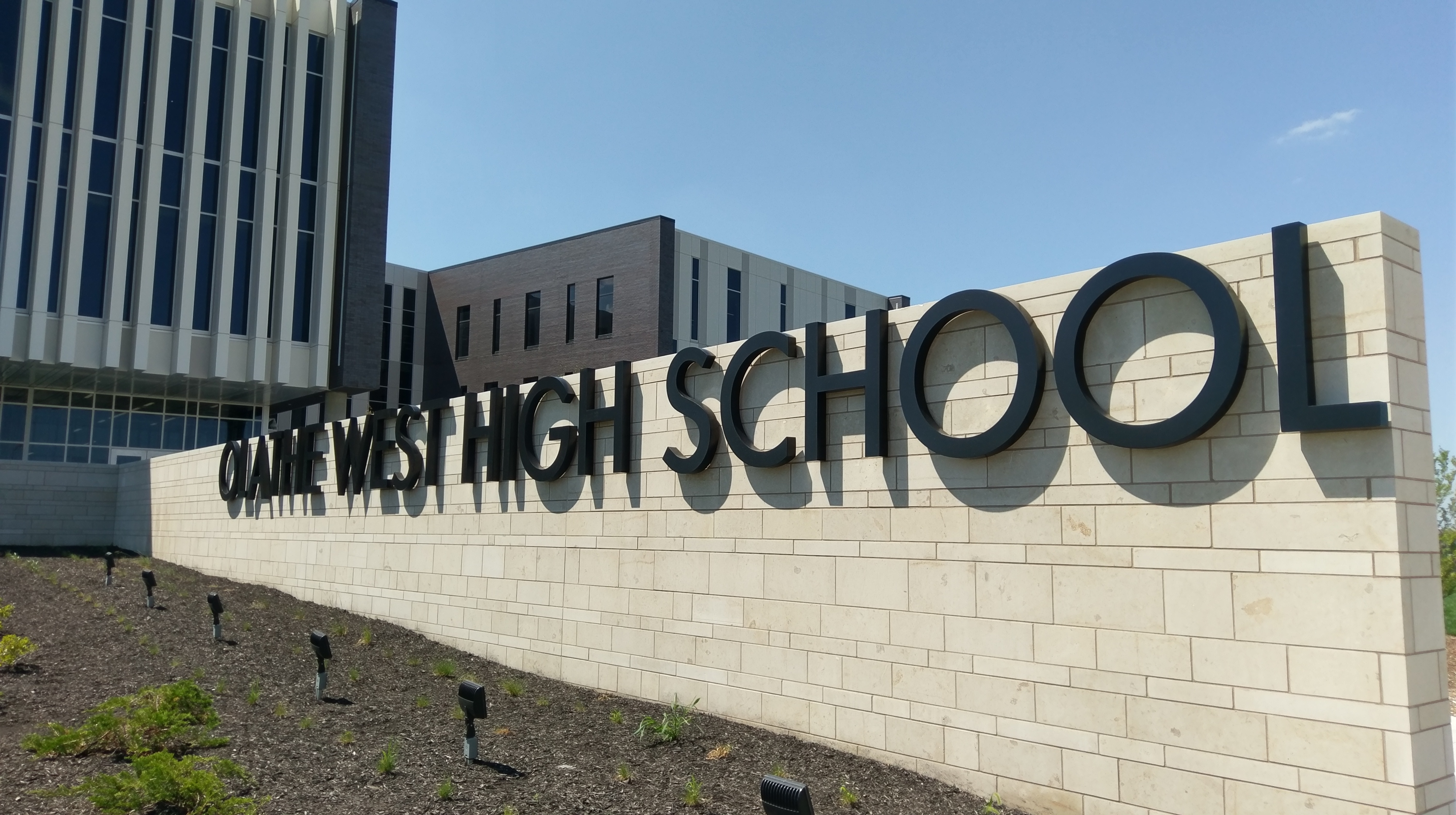 Stone entrance wall reading 'Olathe West High School' with a modern building behind it.