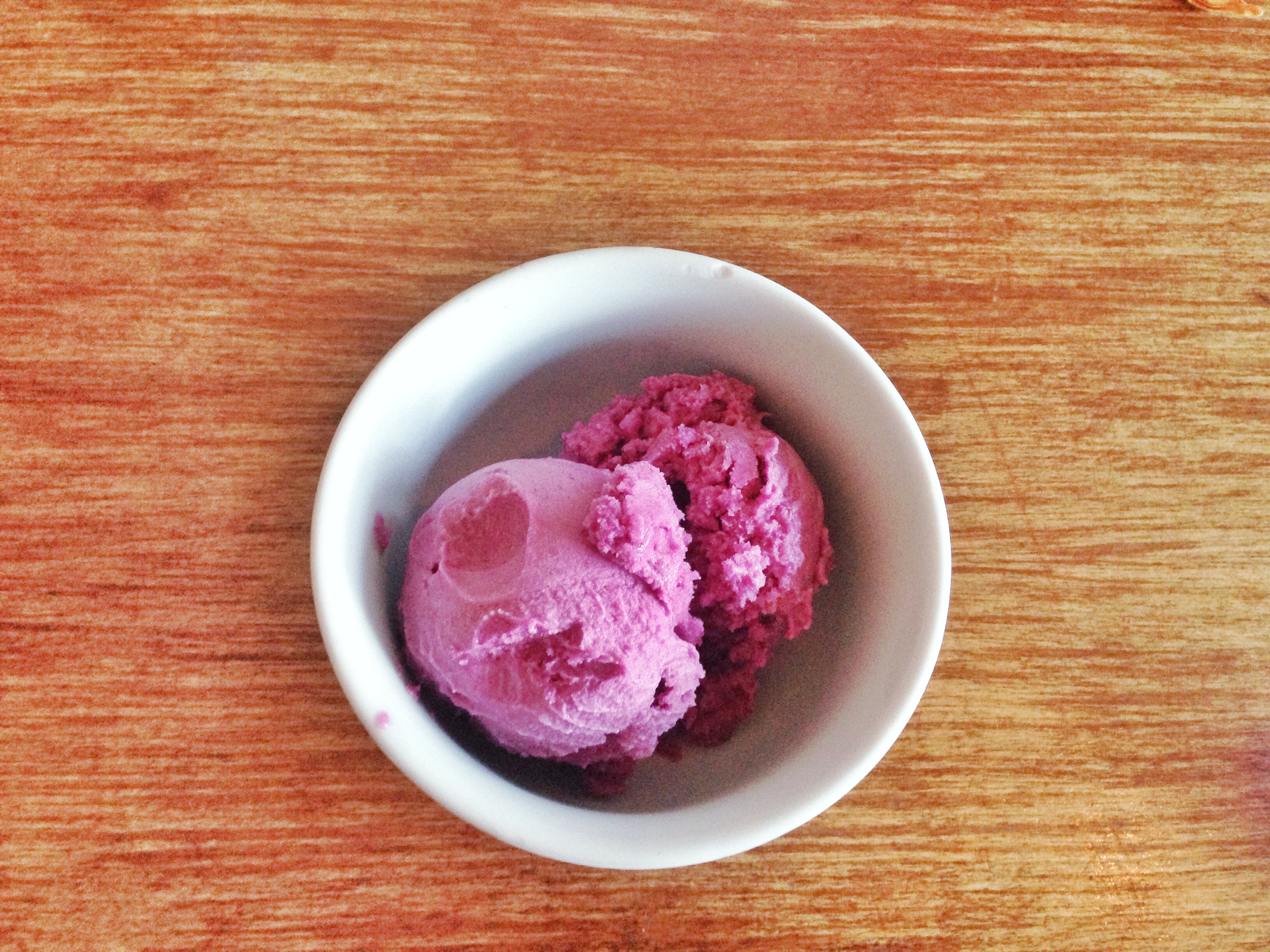 Two scoops of pink-purple sorbet in a small white ceramic bowl on a wooden surface.