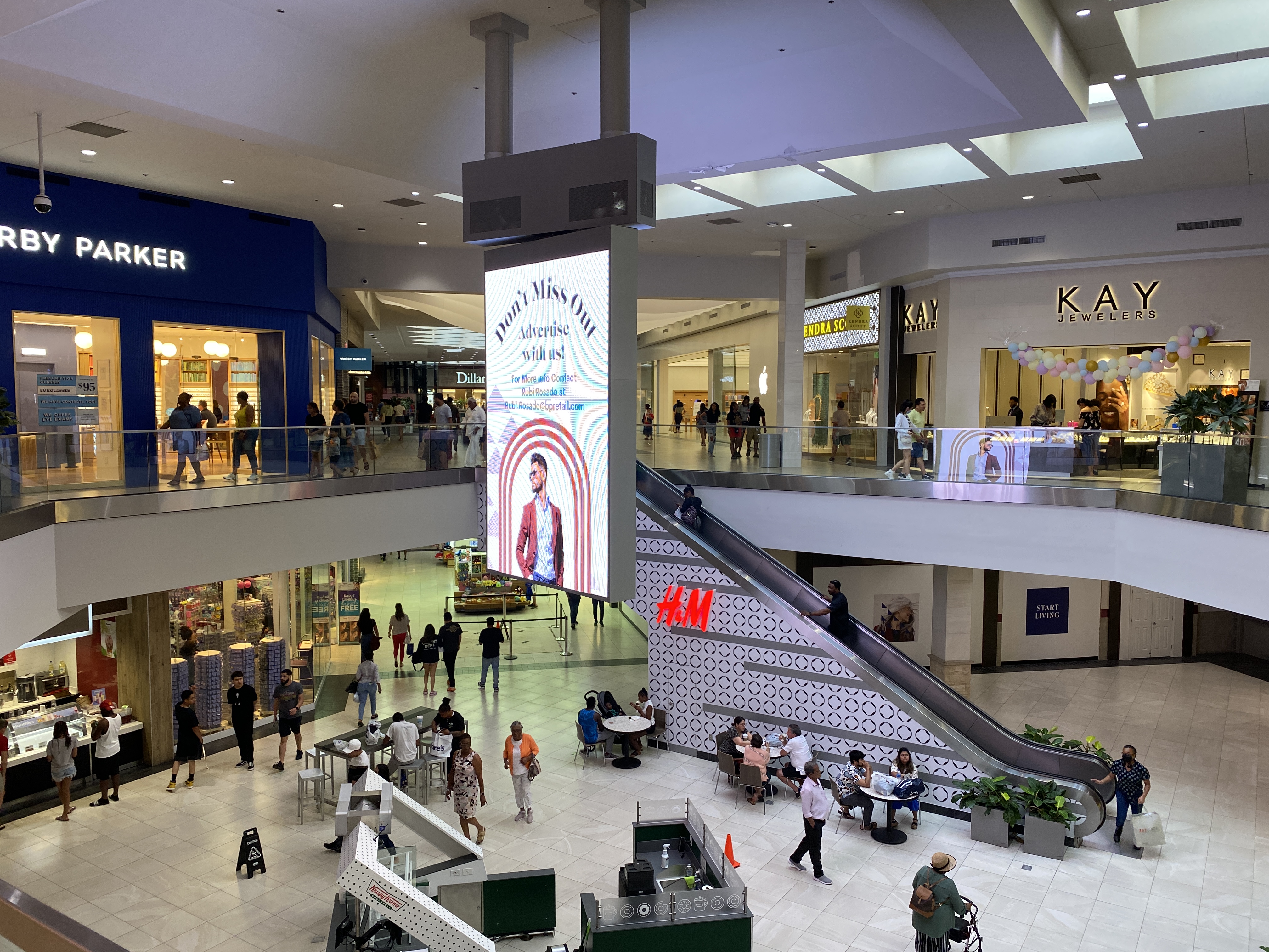 Two-level shopping mall interior with retail stores, escalator, and shoppers.