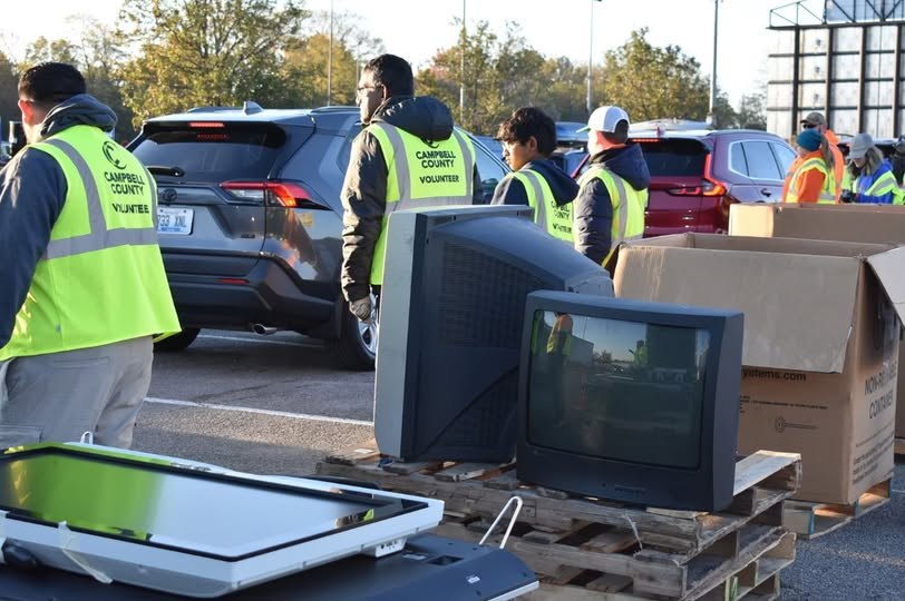 Volunteers in safety vests unload old CRT televisions from vehicles at an outdoor drop-off event.
