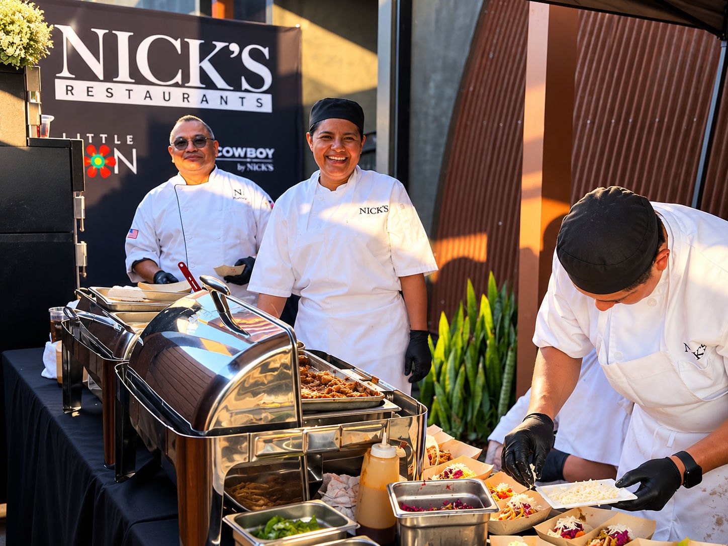 Three chefs in white uniforms serve food from chafing dishes at an outdoor event.