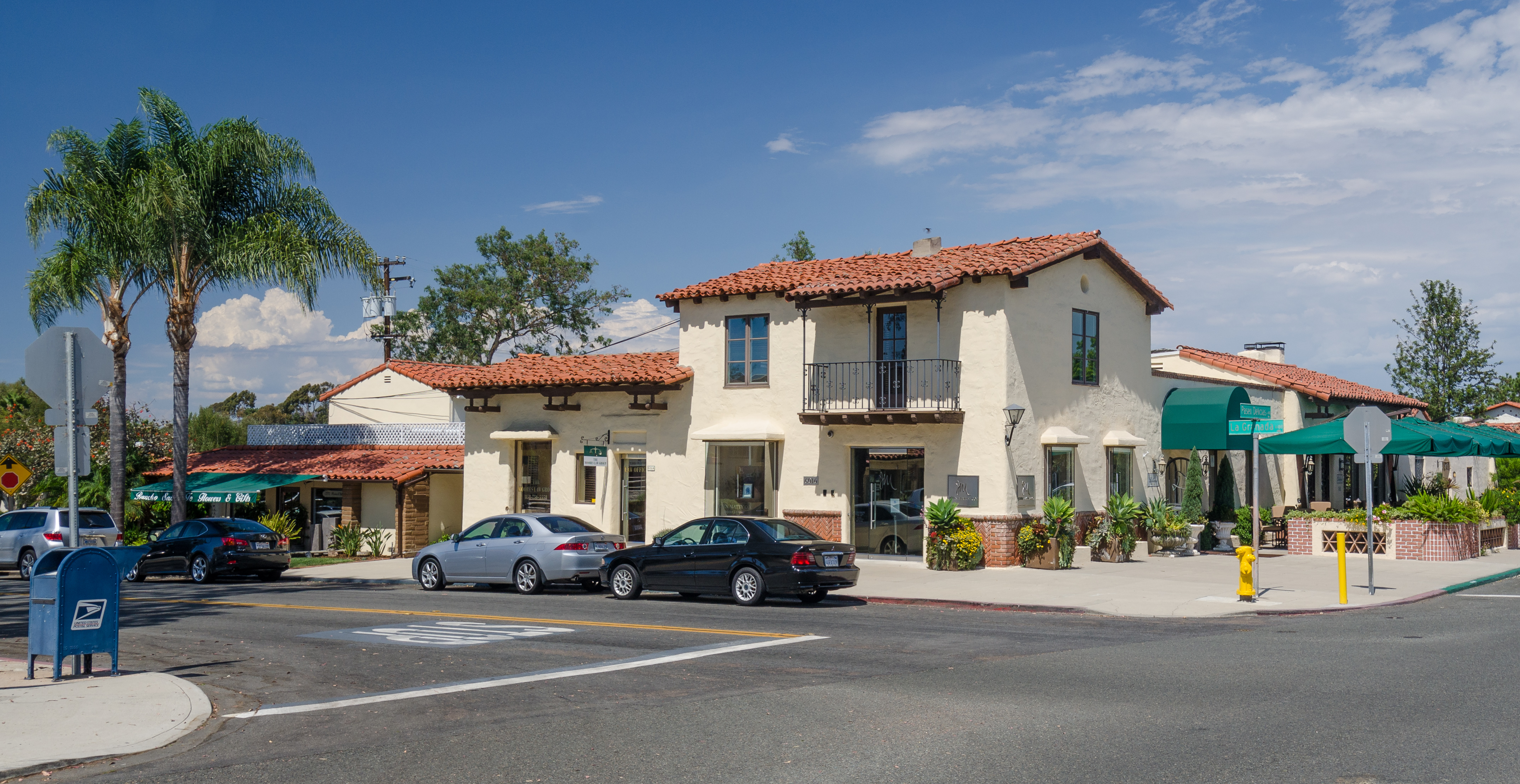 Spanish Colonial Revival building with red tile roof and palm trees on a sunny street.