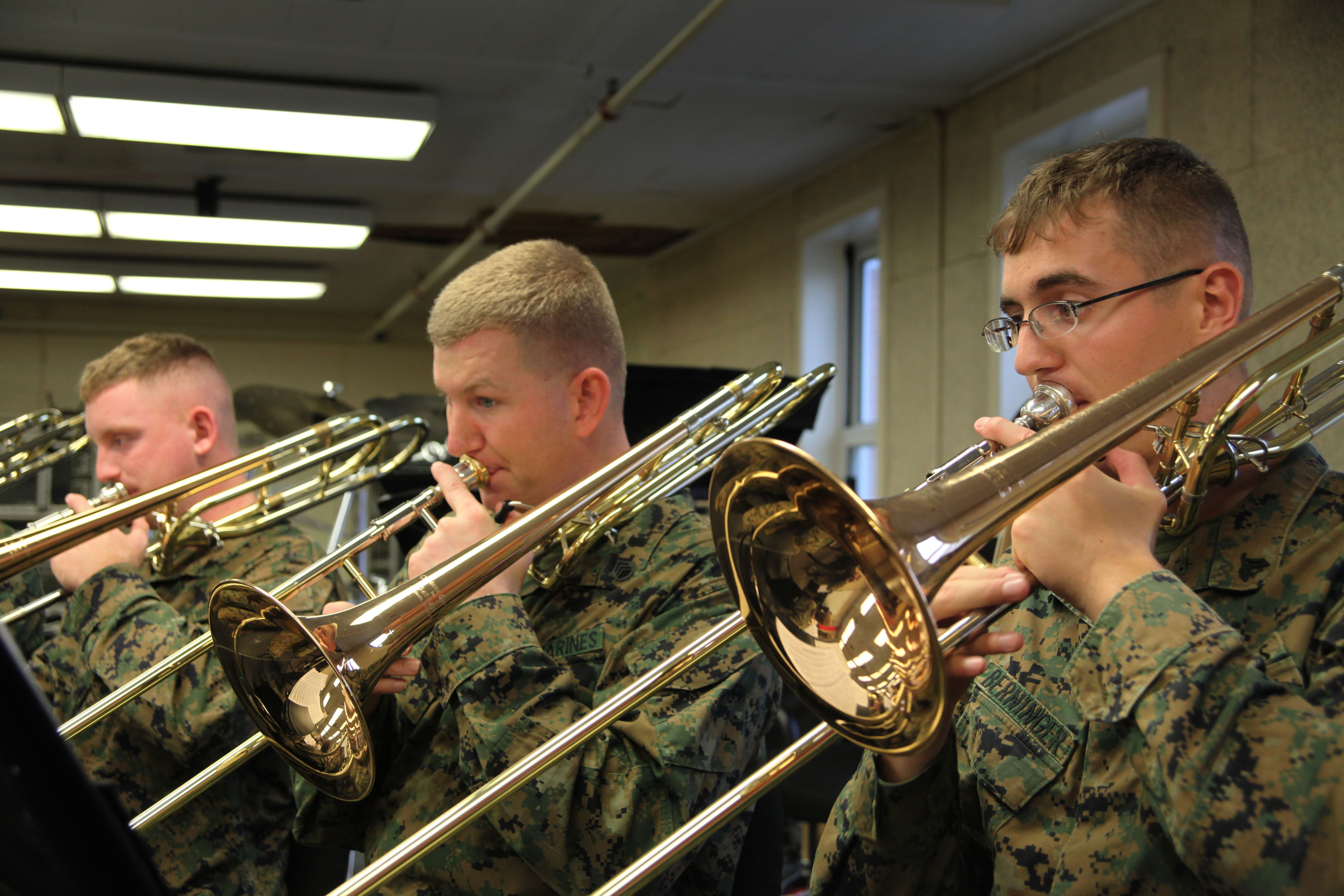 Three Marines in camouflage uniforms playing trombones during an indoor rehearsal.