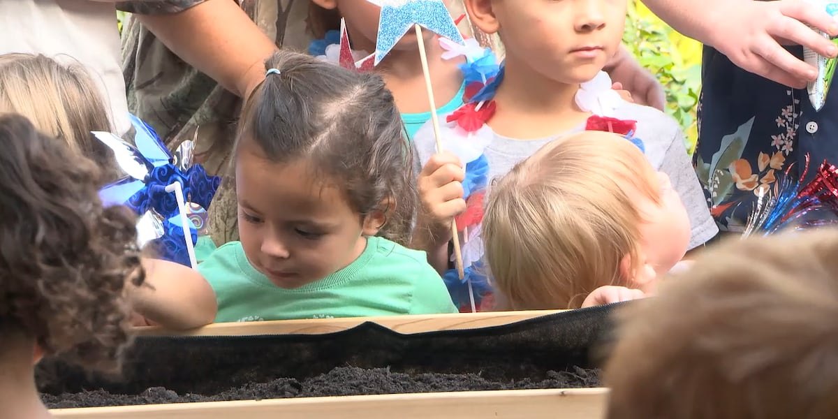Young children crowd around a soil-filled raised garden bed, holding patriotic pinwheels and small American flags.