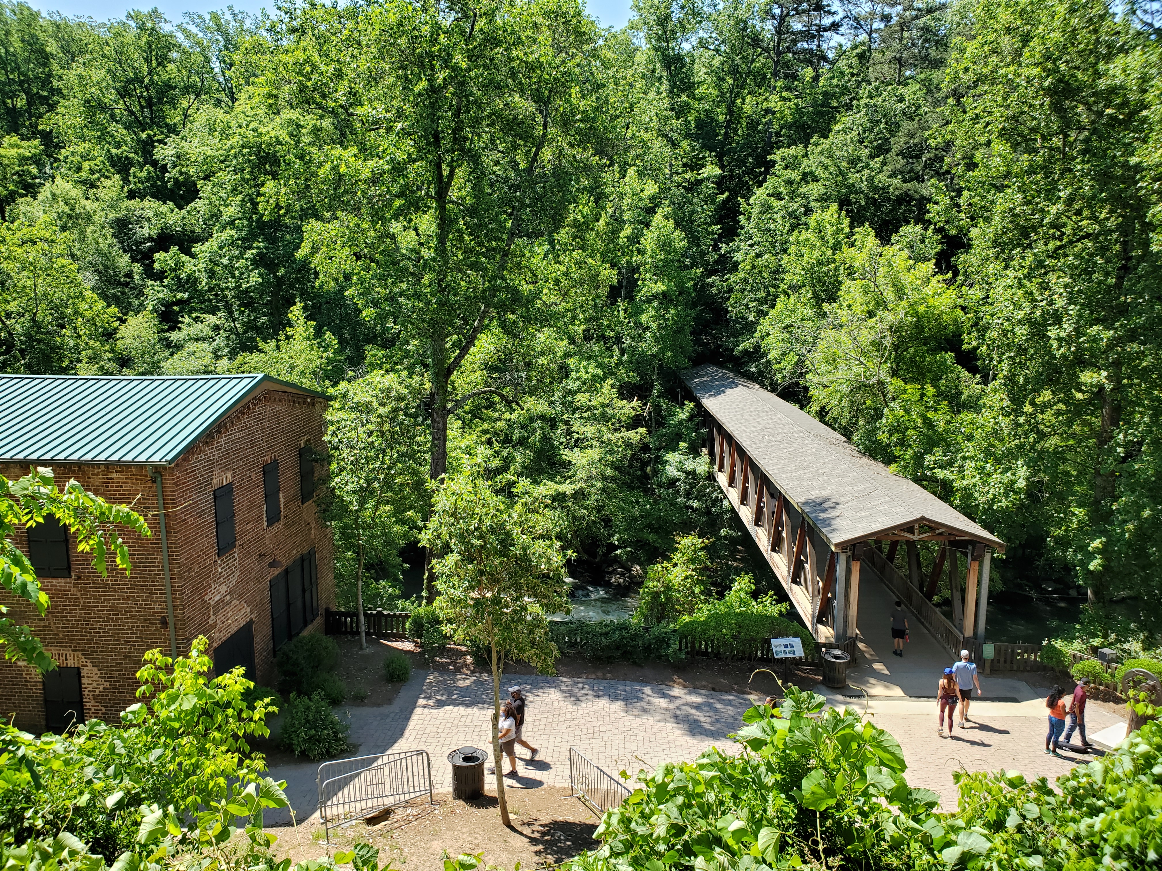 Brick mill building and covered wooden bridge surrounded by dense green forest, with visitors on a gravel path.