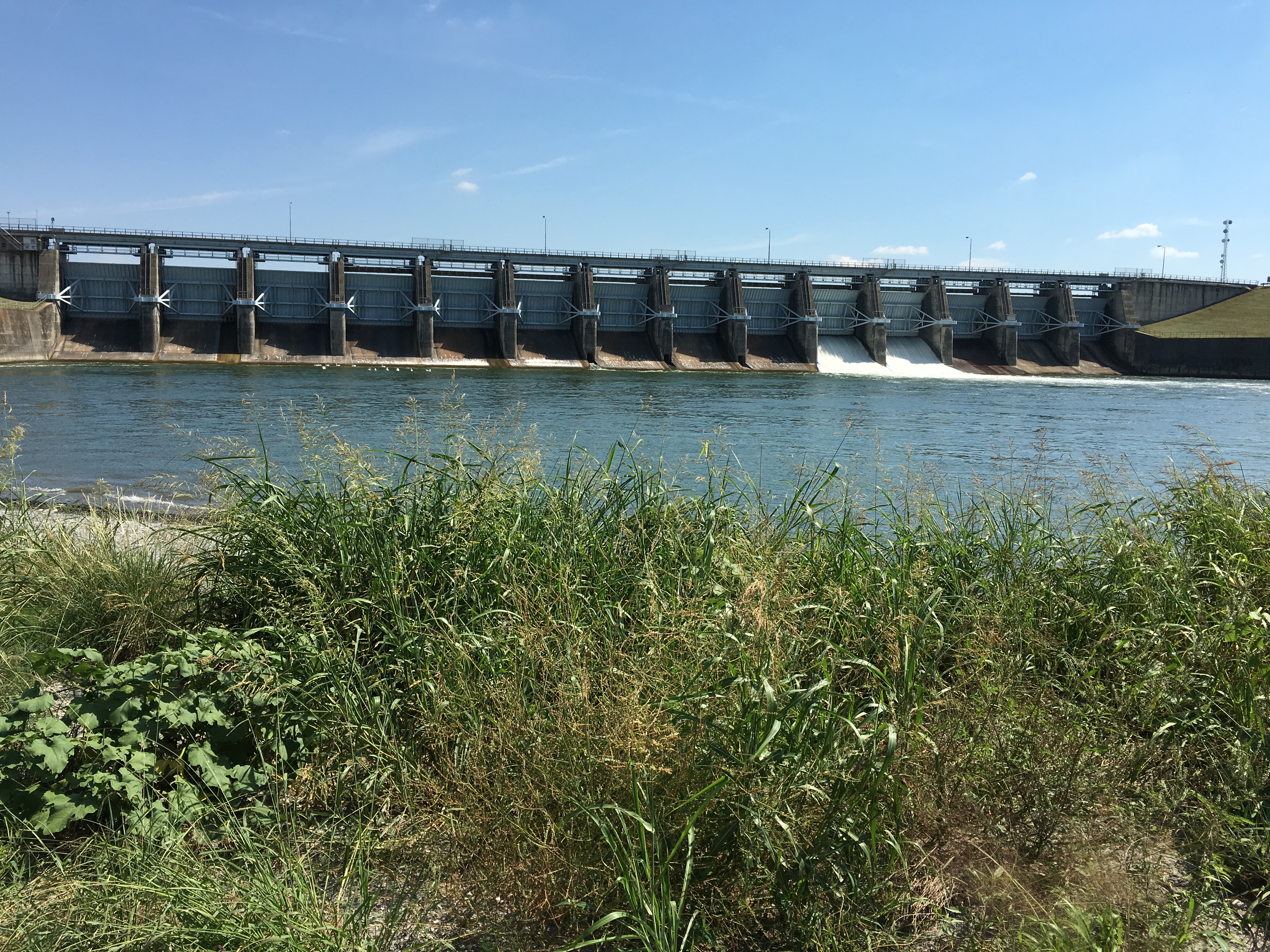 Concrete dam with open spillway gates spanning a wide lake under a clear blue sky.