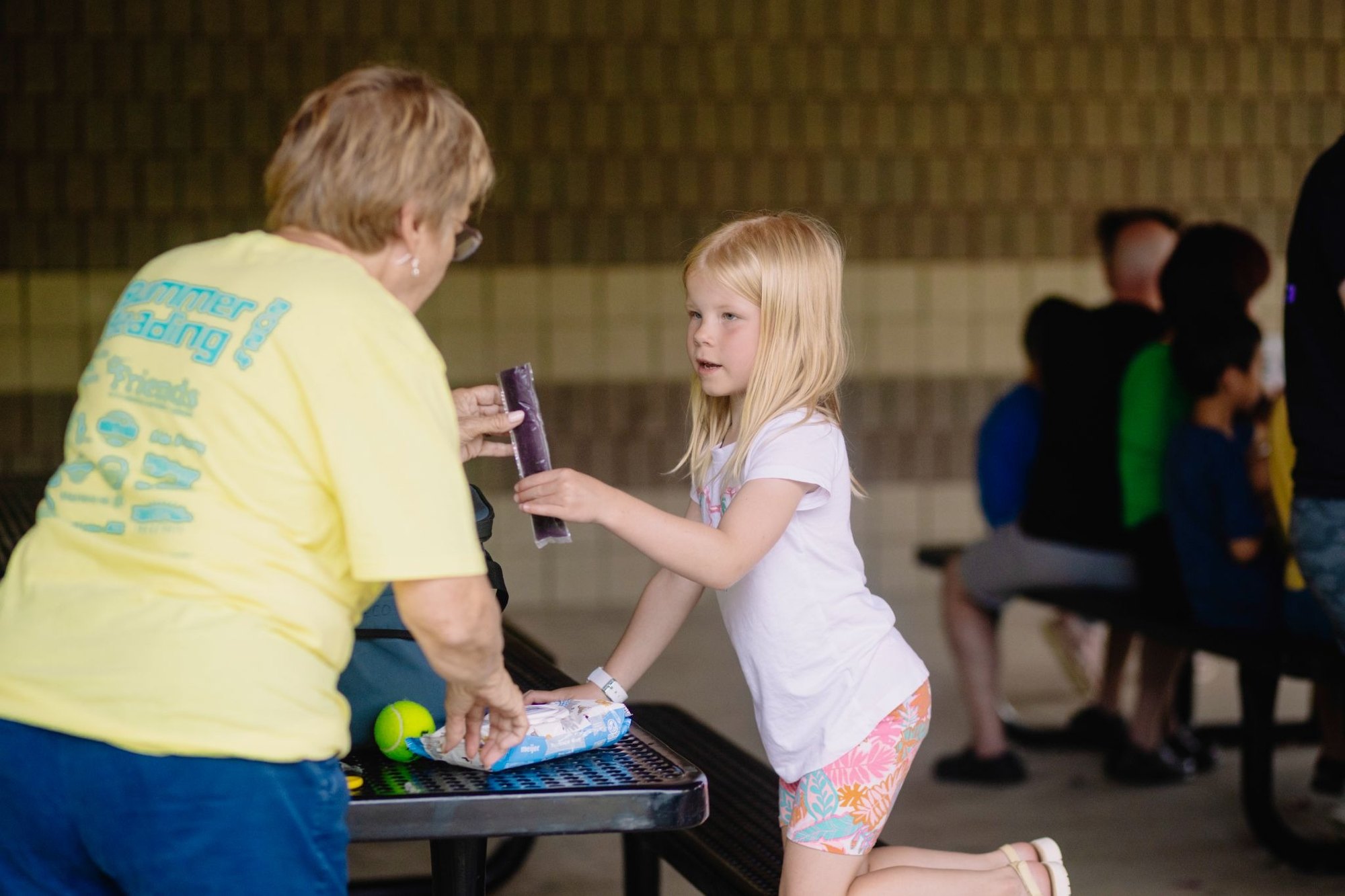A woman in a yellow shirt hands a purple popsicle to a young blonde girl at an outdoor picnic table.